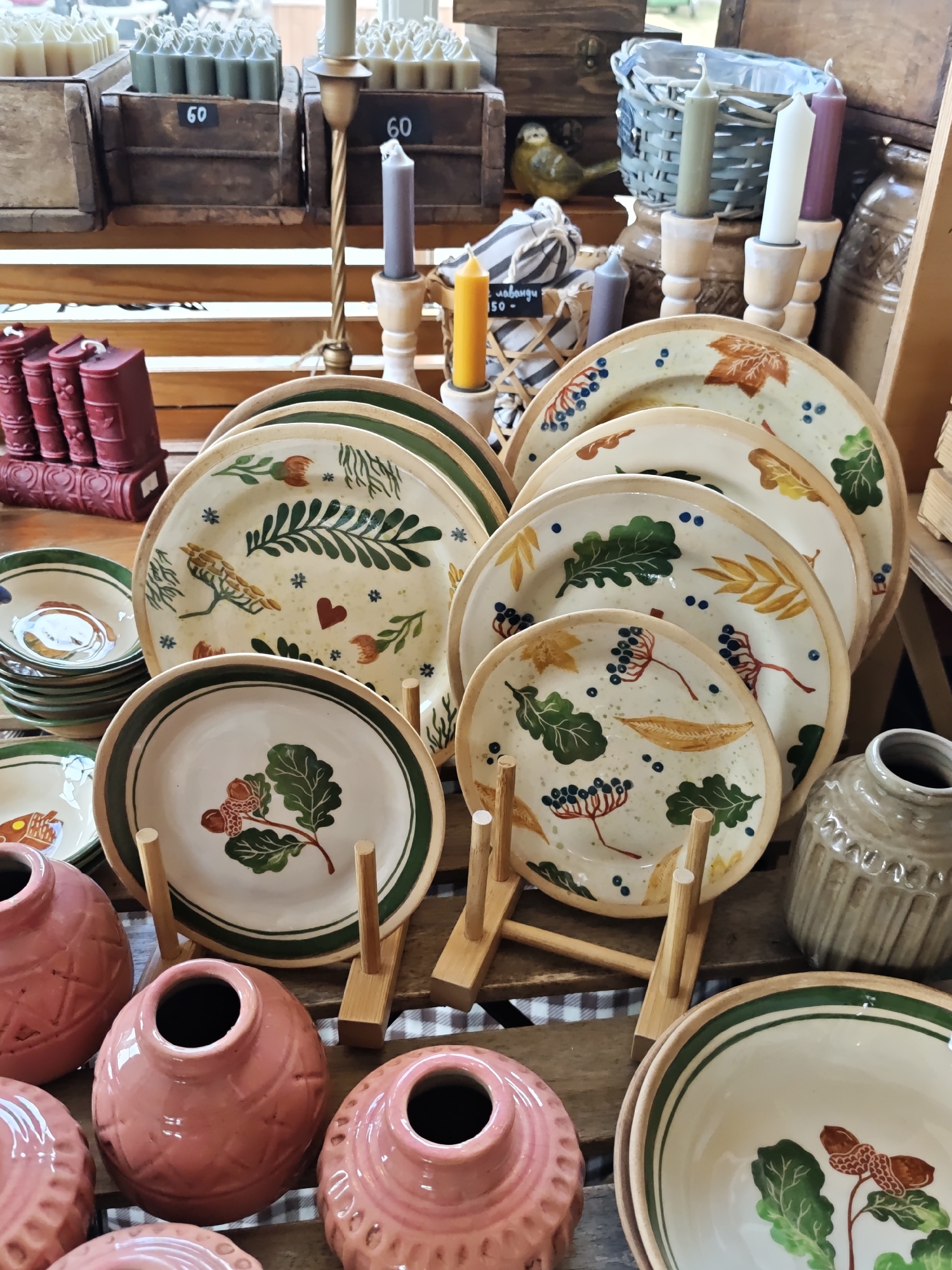 This media file captures a close-up view of a vibrant retail display, likely in a craft shop or market stall in Kyiv, Ukraine. The scene is dominated by an array of handmade ceramic plates and bowls, featuring unique, folk-art style painted designs. These designs include various leaves (oaks, maples), berries, acorns, a small bird, and a red heart, often with green borders. The pottery is displayed on wooden stands and stacked, showcasing a warm, rustic aesthetic.
To the left, a stack of smaller bowls with similar decorative motifs is visible, alongside several ornate, reddish-pink ceramic vases or small pots with textured patterns. In the background and top sections, various candles are arranged. White, grey, green, purple, and yellow candles are placed in wooden or brass holders. Some are neatly arranged in small wooden crates, which bear price tags with the number "60". Another partial tag near a woven basket reads "лаванди - 50-" (likely "lavender - 50"). Other decorative elements include a small, green bird figurine and red, book-shaped objects. The items are arranged on wooden surfaces, some covered with a subtle checkered fabric. The lighting suggests an indoor setting, possibly during the daytime. No people are visible, and the overall mood conveyed is one of charming craftsmanship and cozy home decor.