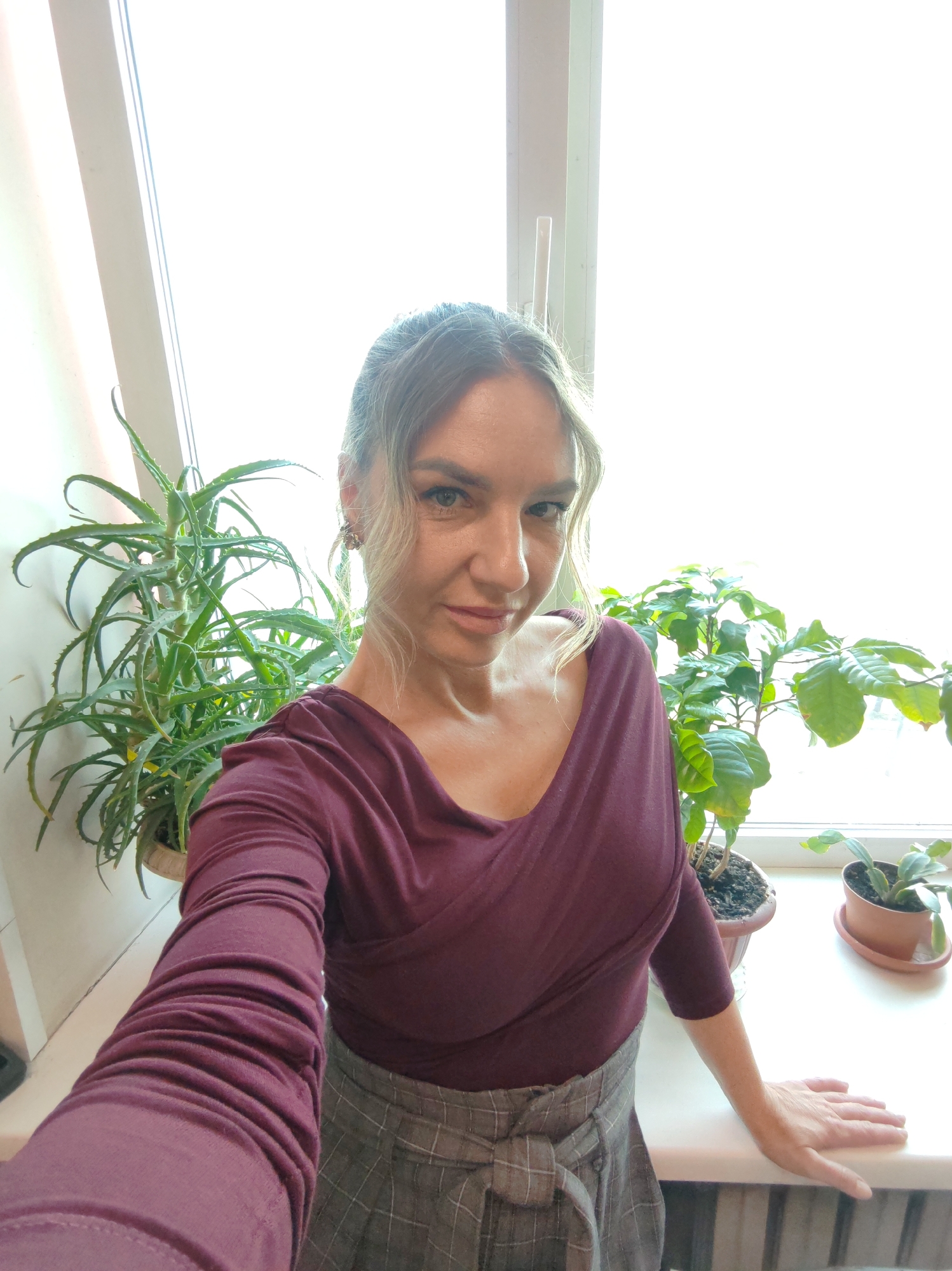 A medium, eye-level shot depicts a woman posing for a selfie in front of a bright window. She has light blonde hair, styled with some strands pulled back, and is wearing a dark maroon, long-sleeved top and a grey, plaid skirt tied at the waist. Her left arm is extended towards the camera, while her right hand rests on the windowsill.
To the woman's left, an aloe plant sits in a light-colored pot. To her right, two potted plants sit on the windowsill. The window itself appears to be a standard double-paned unit.
The scene lacks any distinctive features to determine a specific location. Therefore, it is impossible to confirm if the location is Petropavlovsk-Kamchatsky, Russia, or any other place.