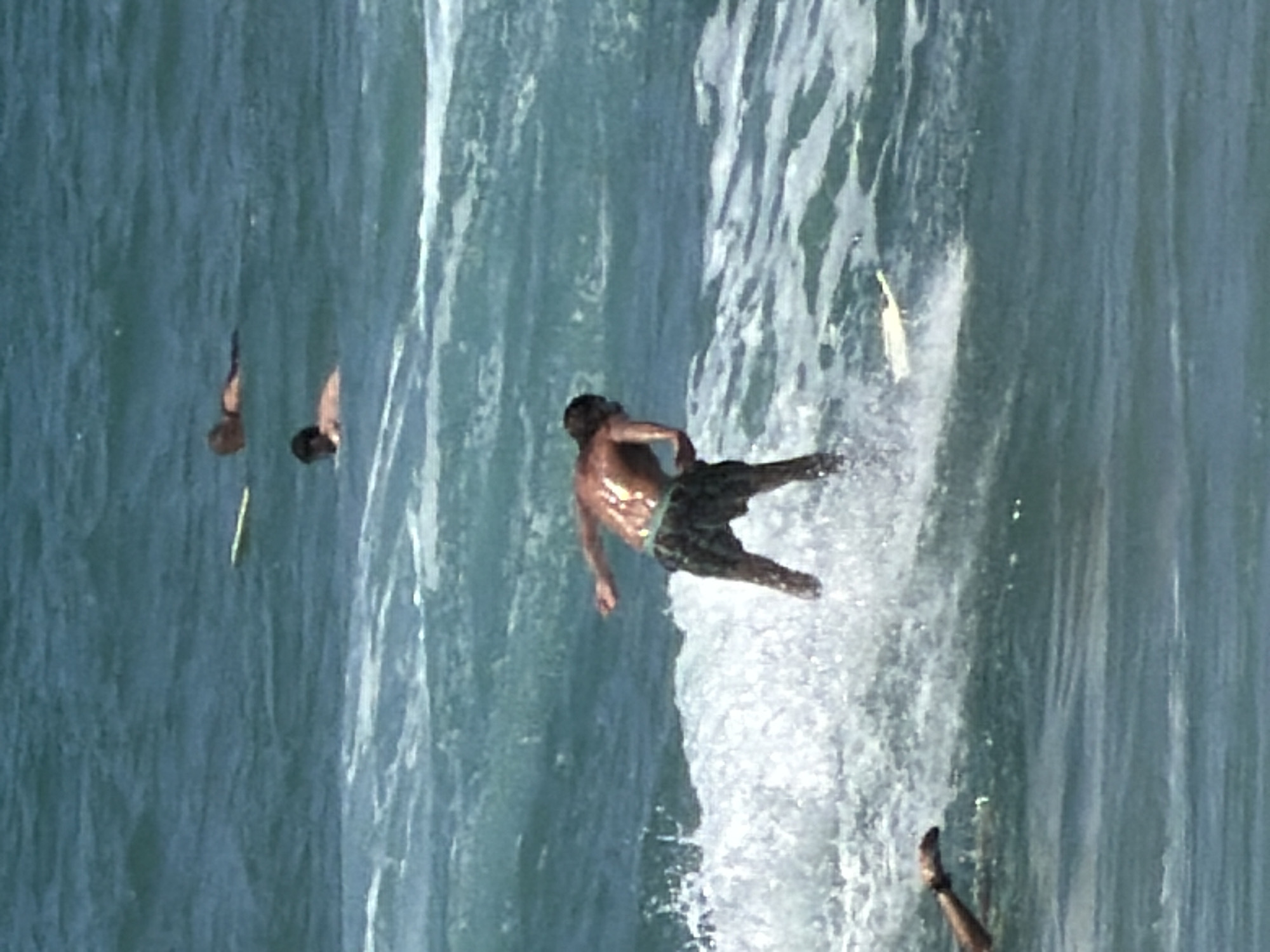 The image shows a group of surfers in the ocean. In the foreground, a surfer in patterned shorts is riding a wave, with their back to the camera. To the left of the frame, another person's arm is visible reaching out of the water. In the background, two other surfers are visible in the water, one lying on a surfboard and another with their head just above the water. The water is choppy and appears to be a deep blue-green color. The lighting suggests a sunny day. The scene is likely taking place at a beach in Tel Aviv, Israel, known for its surfing spots.