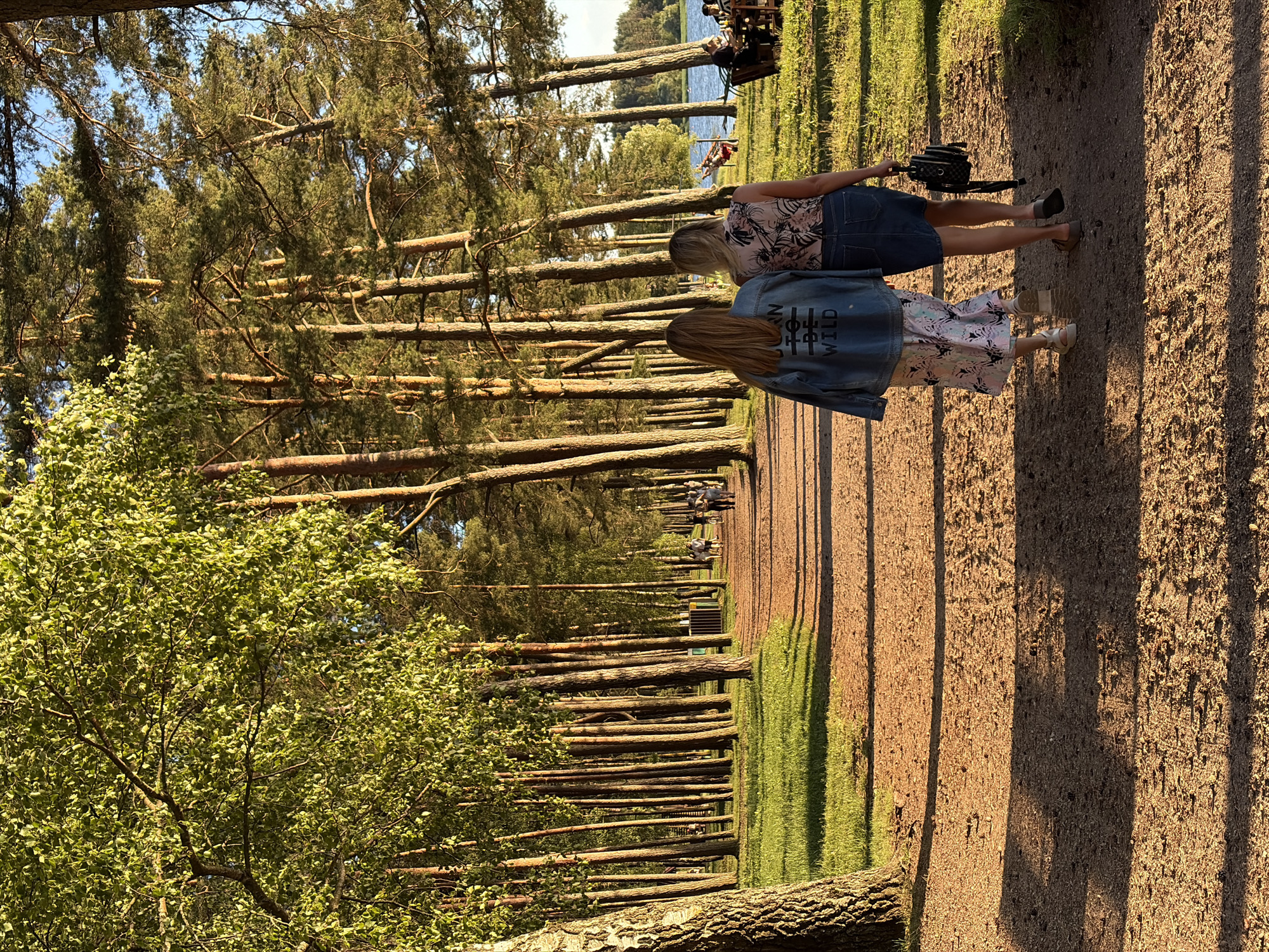 The image depicts a scenic view of a park with a dirt path leading into the distance, lined by tall trees with a sun-dappled forest floor. Two women, viewed from behind, are walking away from the camera on the path. The woman on the left wears a denim jacket with "BORN TO BE WILD" printed on the back, a floral dress, and white sandals. The woman on the right wears a floral print top, a denim skirt, and brown sandals, carrying a black backpack. In the background, several other people are visible, some walking and others seated at tables near a body of water. The environment suggests a park or recreational area with lush green grass and mature trees. The location is identified as Ждановіцкі сельскі Савет, Belarus.