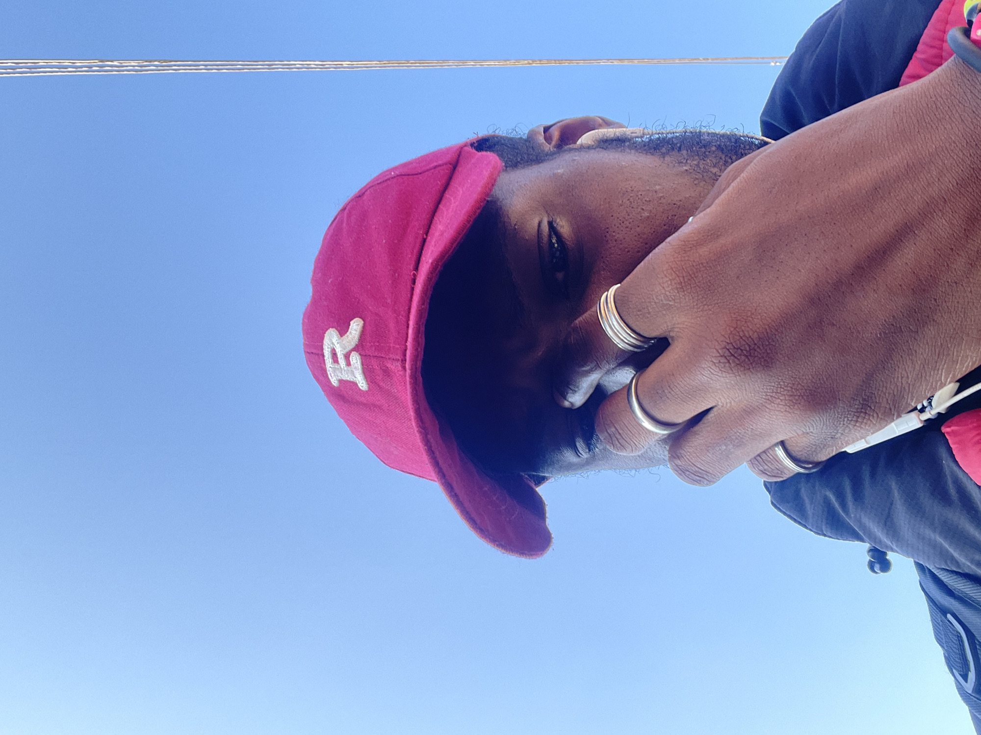 A close-up, low-angle outdoor shot shows a person wearing a bright pink baseball cap with a white "R" logo. The person's face is partially obscured by their hand, which is adorned with multiple silver rings. The background is a clear, bright blue sky, suggesting it is daytime. A thin, vertical element, possibly a cable or antenna, is visible on the far right of the frame. The image does not contain any specific landmarks or visual cues that would allow for identification of the location as Jos, Nigeria.