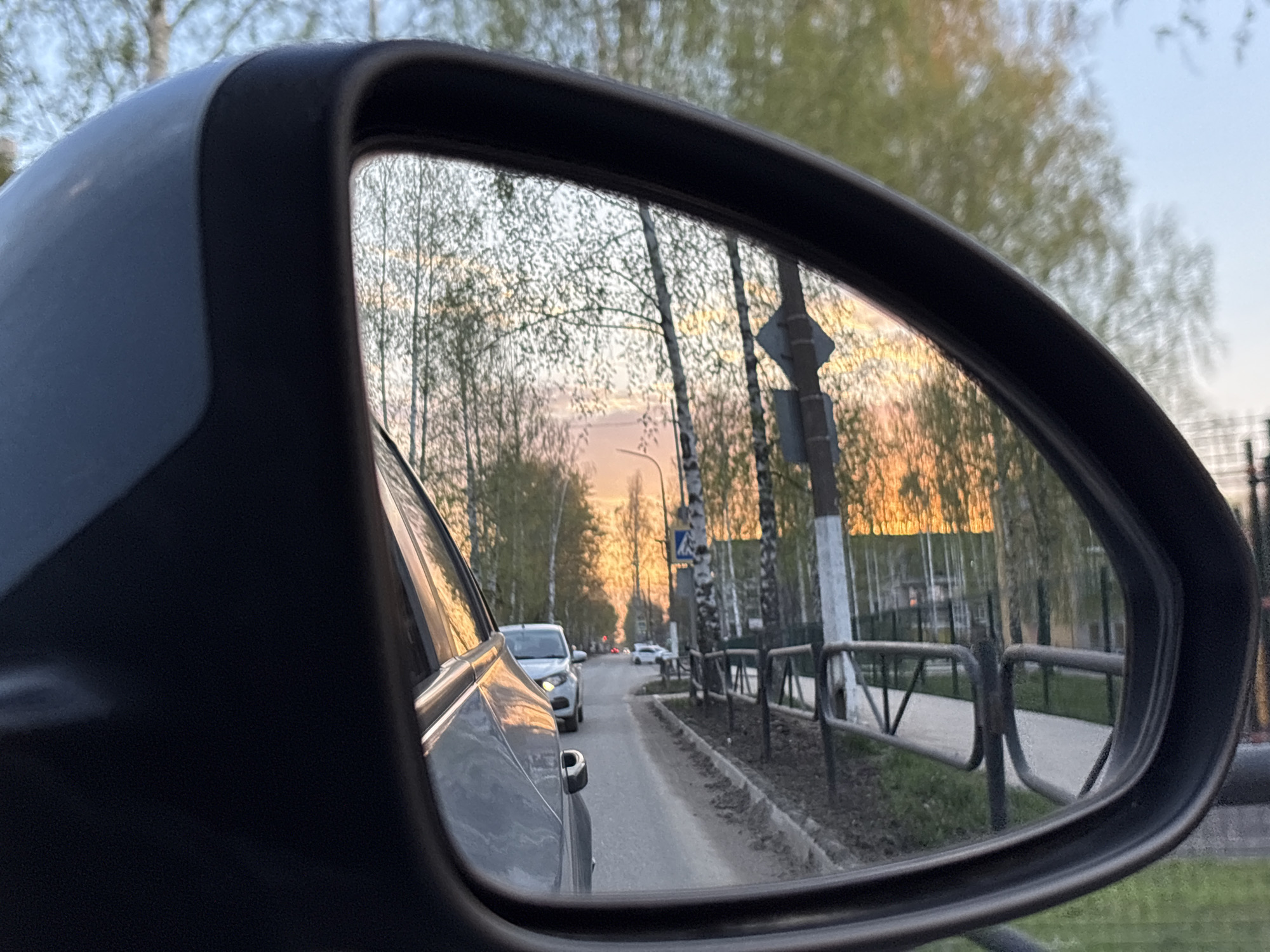 The image shows a view through a car's side mirror. In the reflection, a street stretches into the distance with trees lining both sides. The sky is a gradient of orange and light blue, suggesting either sunrise or sunset. There are several cars on the street, including a white car closer to the viewer. A pedestrian walkway with a metal fence runs alongside the road. A sign is visible, mounted on a pole with white and gray paint. The environment appears to be a residential area, as suggested by the fences. The location is a street in a city.