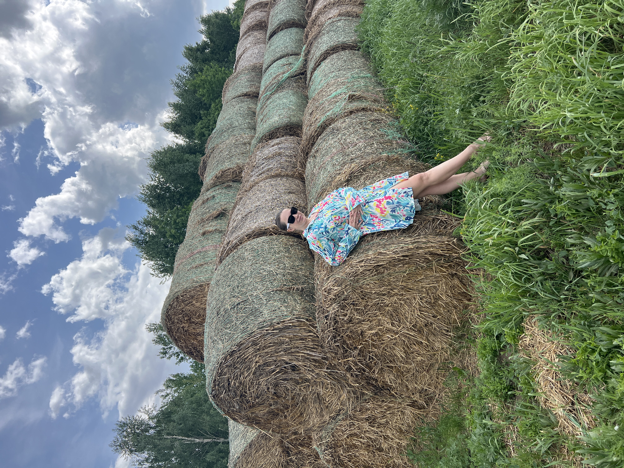 A woman wearing a colorful dress and sunglasses lies on a stack of hay bales in a field. The hay bales are stacked in multiple rows, extending upwards towards the top of the frame. Green grass and vegetation border the hay bales on one side. Trees are visible in the background along with a sky filled with clouds. The location is a rural area, potentially near Kirillovka, Russia. The woman appears to be posing for a photograph.