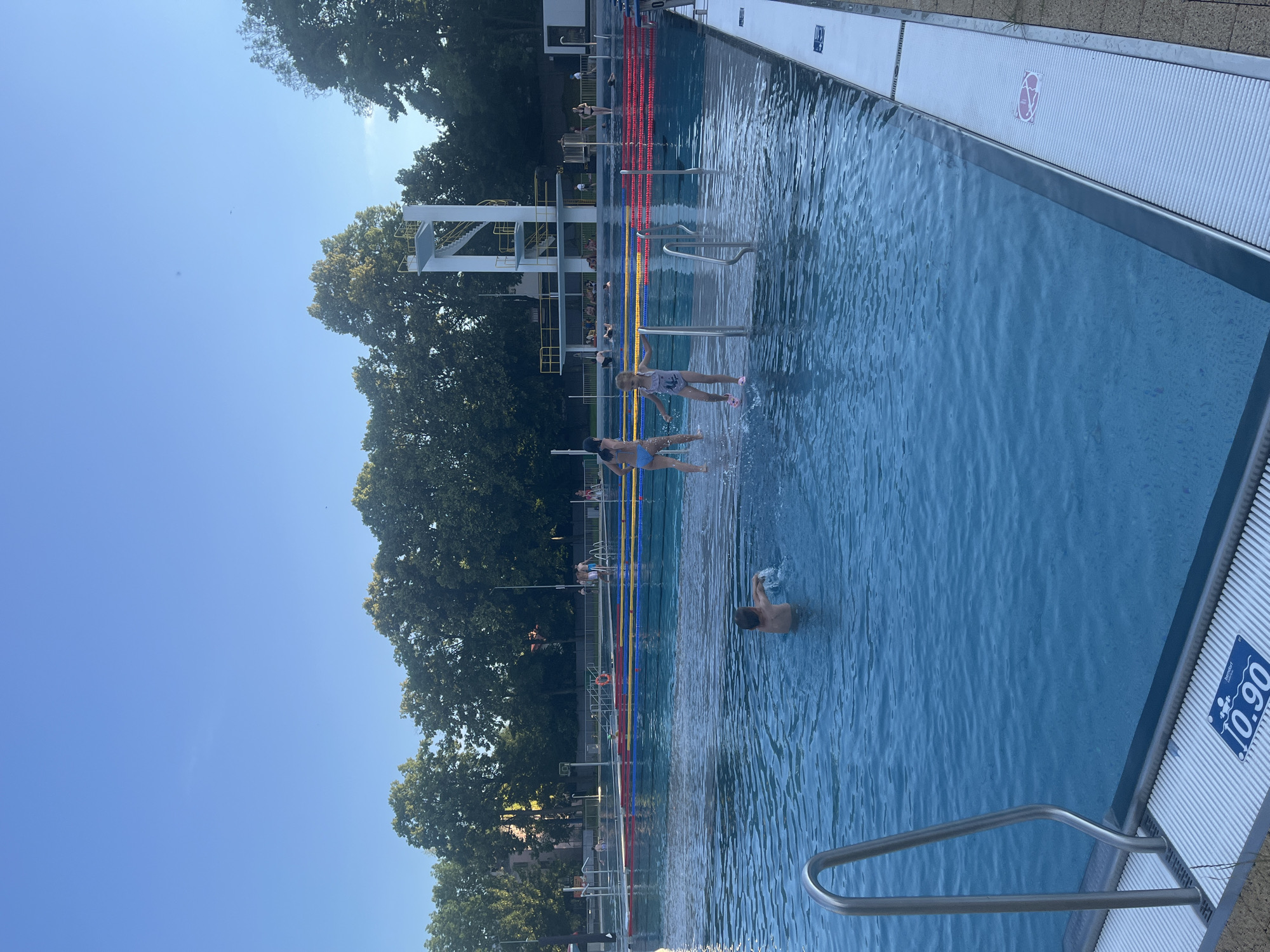 The image depicts an outdoor swimming pool on a clear day. In the foreground, a young boy splashes water. Further back, two young girls in swimwear are playing in the shallow end of the pool. The pool is marked with lanes and has a diving board structure in the background. Several other people are visible in the pool and around its edges, some resting or observing. Lush green trees line the perimeter of the pool area, suggesting a park-like setting. A sign at the edge of the pool indicates a depth of 0.90. The sky is a clear blue.