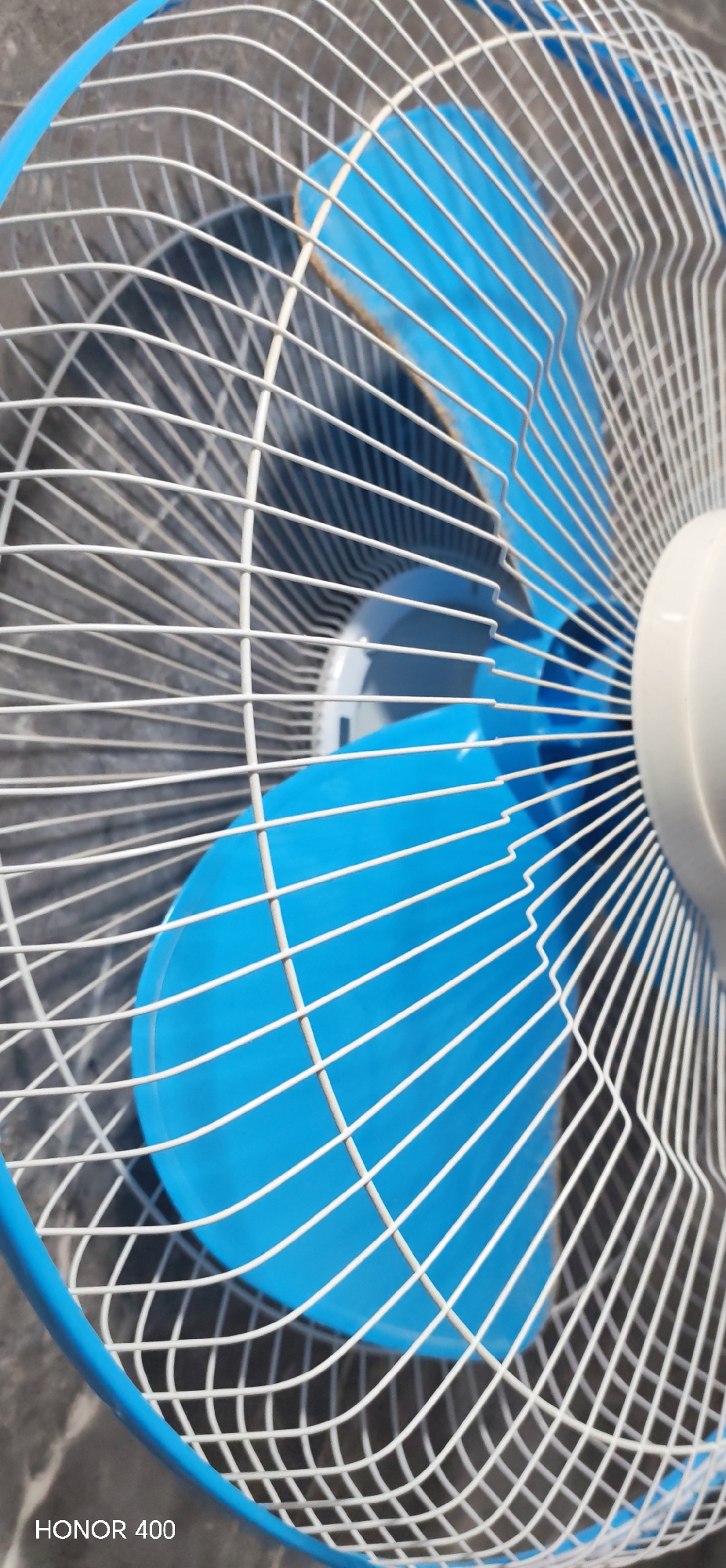 A close-up shot features a blue electric fan behind a white wire cage. The fan is partially visible, with its blades angled towards the viewer. The backdrop consists of a blurred, grey-toned surface. The words "HONOR 400" are visible in the lower left corner.