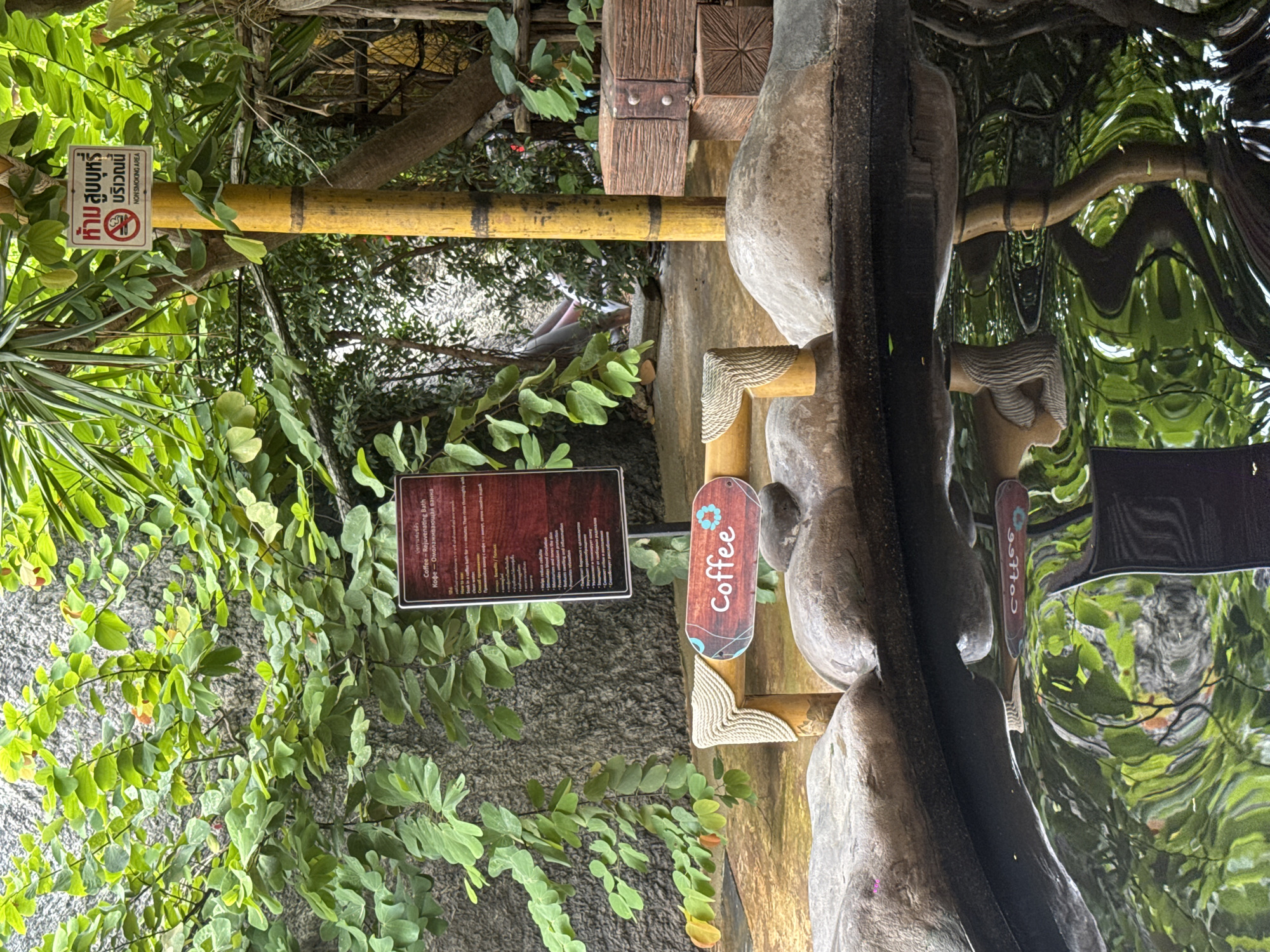 The image captures an outdoor scene in Ban Khlong Khian, Thailand, featuring signs related to coffee and smoking regulations. On the left, lush green vegetation frames a sign that provides information on a "Coffee - Rejuvenating Bath," with the text also translated into Russian. Adjacent to this, there is a "Coffee" sign, written vertically. A yellow bamboo pole bears a "No Smoking" sign, indicating a designated non-smoking area. The backdrop includes a weathered gray surface and reflections suggesting a body of water. The overall setting appears to be a garden or outdoor area associated with a spa or wellness facility.