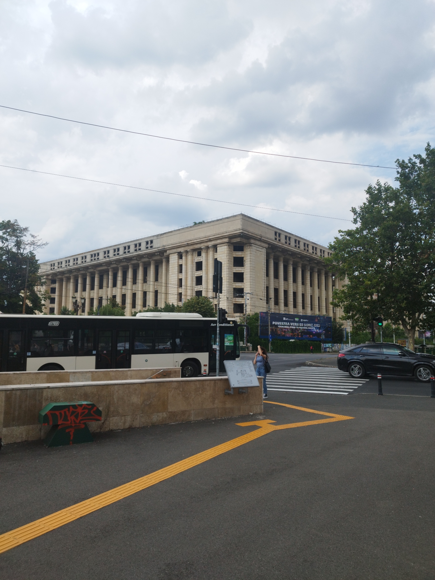 The image captures a street scene in Bucharest, Romania, with a large, classical-style building dominating the background. The building features a long facade with numerous columns and arched windows. In the foreground, a white and black bus is stopped, possibly at a bus stop. A person is standing on the sidewalk next to the bus, looking at their phone. A black car is visible on the street, crossing a pedestrian crosswalk. The street is paved with asphalt, and there are yellow tactile paving lines on the sidewalk. The sky is cloudy, suggesting overcast weather. A large billboard with text in Romanian is mounted on the side of the building. The overall impression is a typical urban environment with elements of public transportation, architecture, and pedestrian activity.