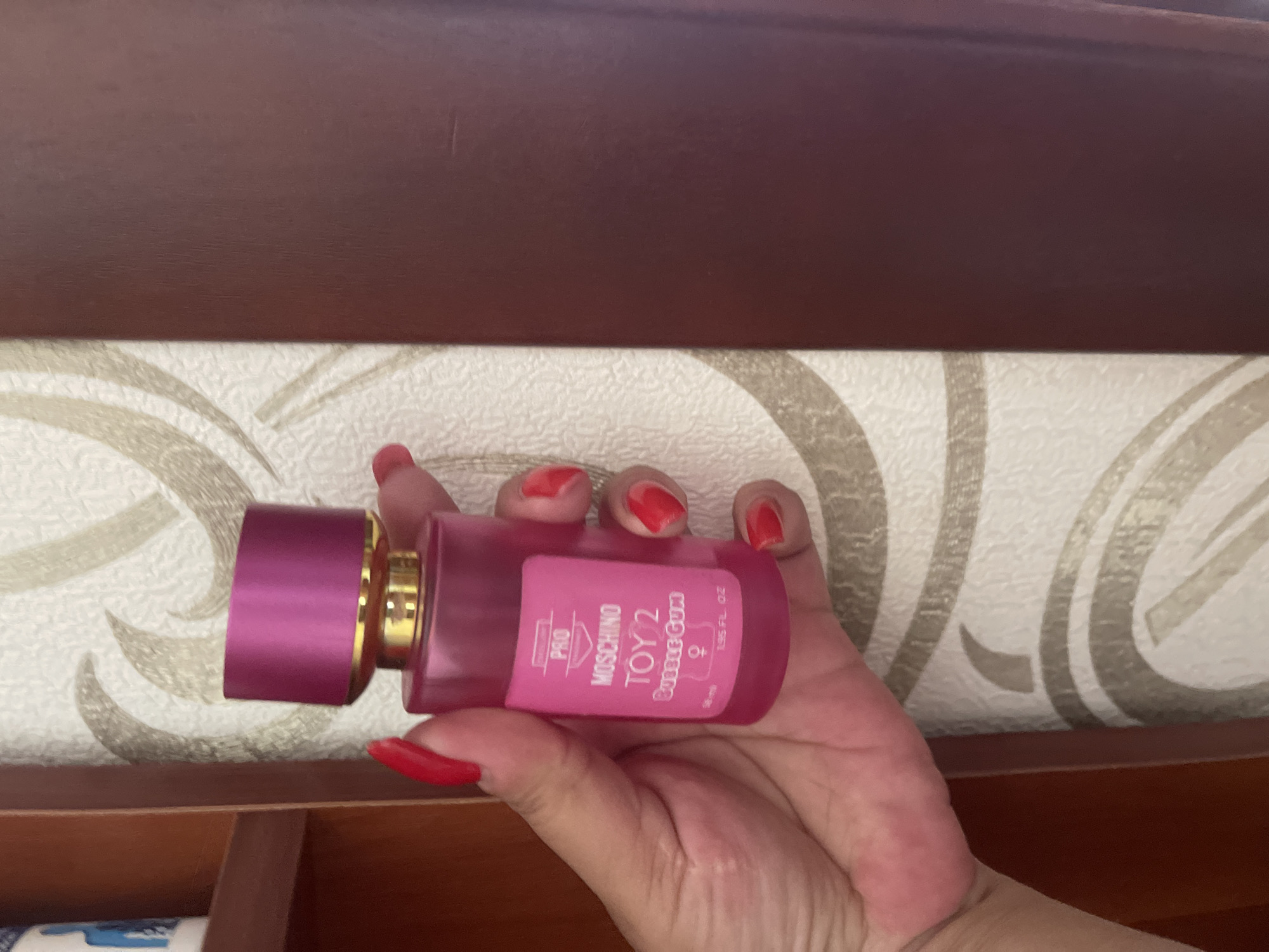 A person is holding a pink bottle of Moschino Toy 2 Bubble Gum fragrance. The bottle has a gold accent and a pink cap. The background consists of wallpaper with a swirling pattern and a dark brown surface. The scene is indoors.