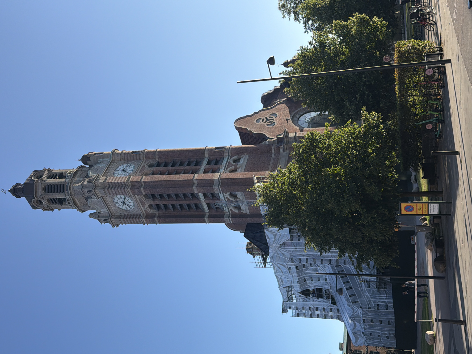 The image captures a street view of the Caroli Church in Malmö, Sweden, under renovation. A tall, brick clock tower dominates the right side of the frame, featuring ornate architectural details and a cross at its apex. The main body of the church, to the left of the tower, is partially obscured by scaffolding and white tarpaulin, indicating ongoing construction work. Lush green trees are in the foreground, with a paved area and a sidewalk in front of the church. Several parked scooters and bicycles are visible on the right side of the sidewalk, near a hedge. Two individuals are standing near a barrier in front of the construction-covered portion of the church. The sky is clear and blue, suggesting fair weather.