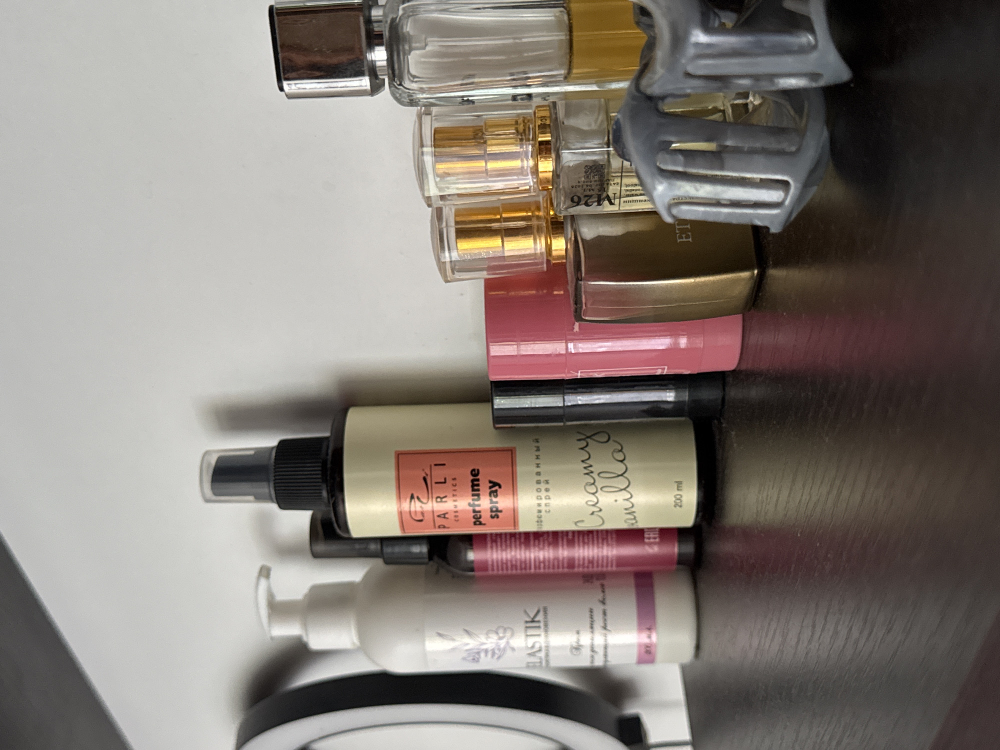 A collection of personal care products and perfumes are arranged on a dark wooden shelf against a white background. From left to right, a white pump bottle with a purple label is visible, followed by a tall, thin pink bottle. Next is a larger, light green bottle with an orange label that reads "PARLI COSMETICS perfume spray Creamy vanilla 200 ml". To the right of this are several small perfume bottles with gold accents and clear caps, and a larger glass bottle containing a yellow liquid. A grey plastic object, possibly a makeup tool or accessory, is in the foreground. The scene is lit by soft light, casting subtle shadows. The overall impression is one of personal grooming items organized on a shelf. The context of Яромінскі сельскі Савет, Belarus, is not visually represented in the image.