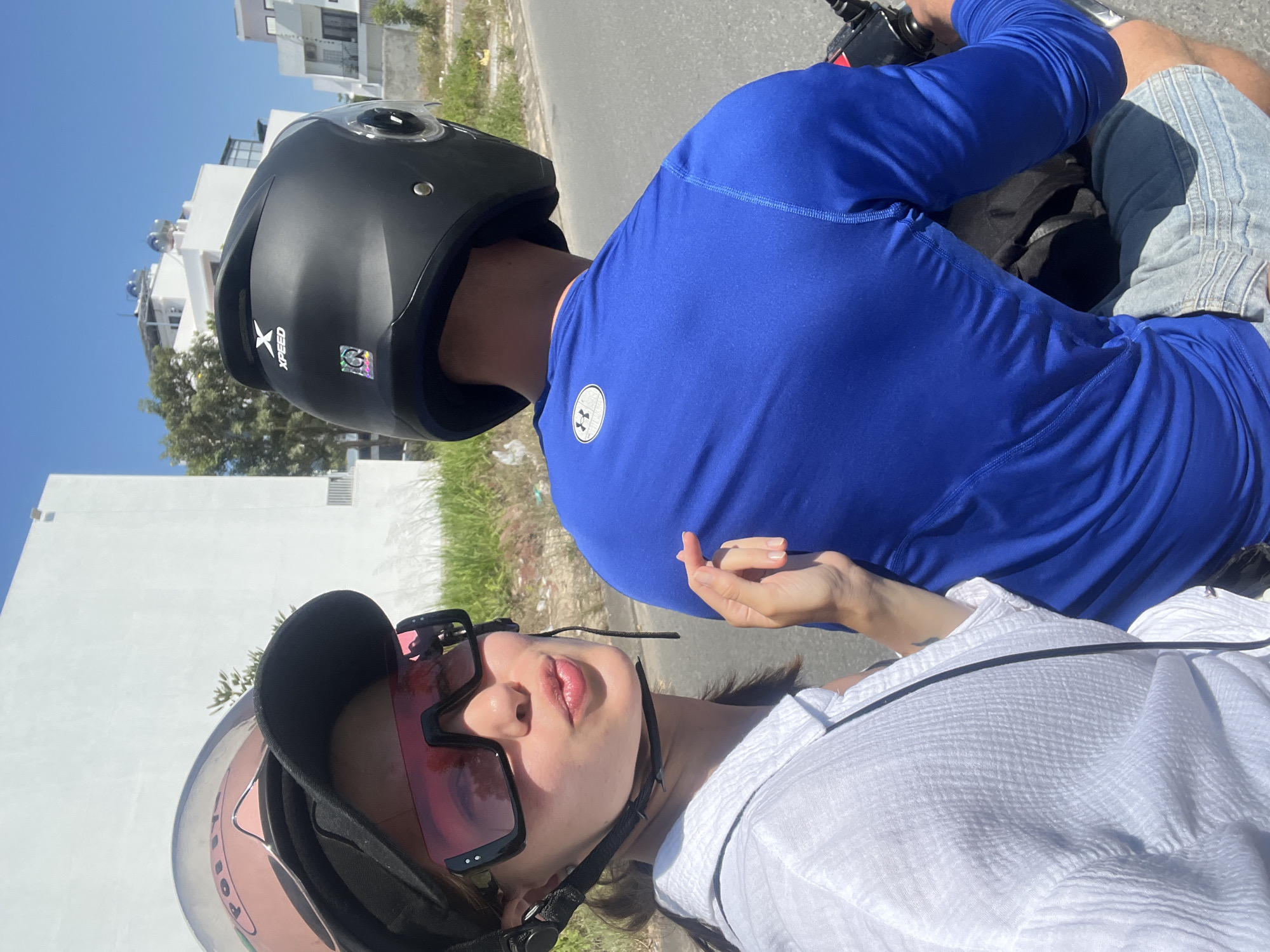 Two individuals are depicted on a motorcycle in Nha Trang, Vietnam, under a clear blue sky. The person in the foreground, a female, occupies the passenger position and faces the camera, making a pouting expression with her lips. She wears a white textured long-sleeved shirt, a black baseball cap, and oversized black-framed sunglasses with pink-tinted lenses. A pink-tinted helmet is visible over her cap, reflecting "POLICE" lettering. Her right hand is raised, with fingers slightly curled, positioned near the driver's back. The driver, a male, is seated in front, facing away from the camera. He wears a royal blue long-sleeved athletic shirt with a white Under Armour logo on the upper back and light blue denim shorts. He wears a black open-face helmet with an "XPEED" logo on the rear. The motorcycle handlebars are partially visible. The background features several white, multi-story buildings with flat roofs, some displaying water heating systems or tanks. Sparse green vegetation, including grass and a small tree, lines the edge of a paved road. The scene is brightly lit by direct sunlight.