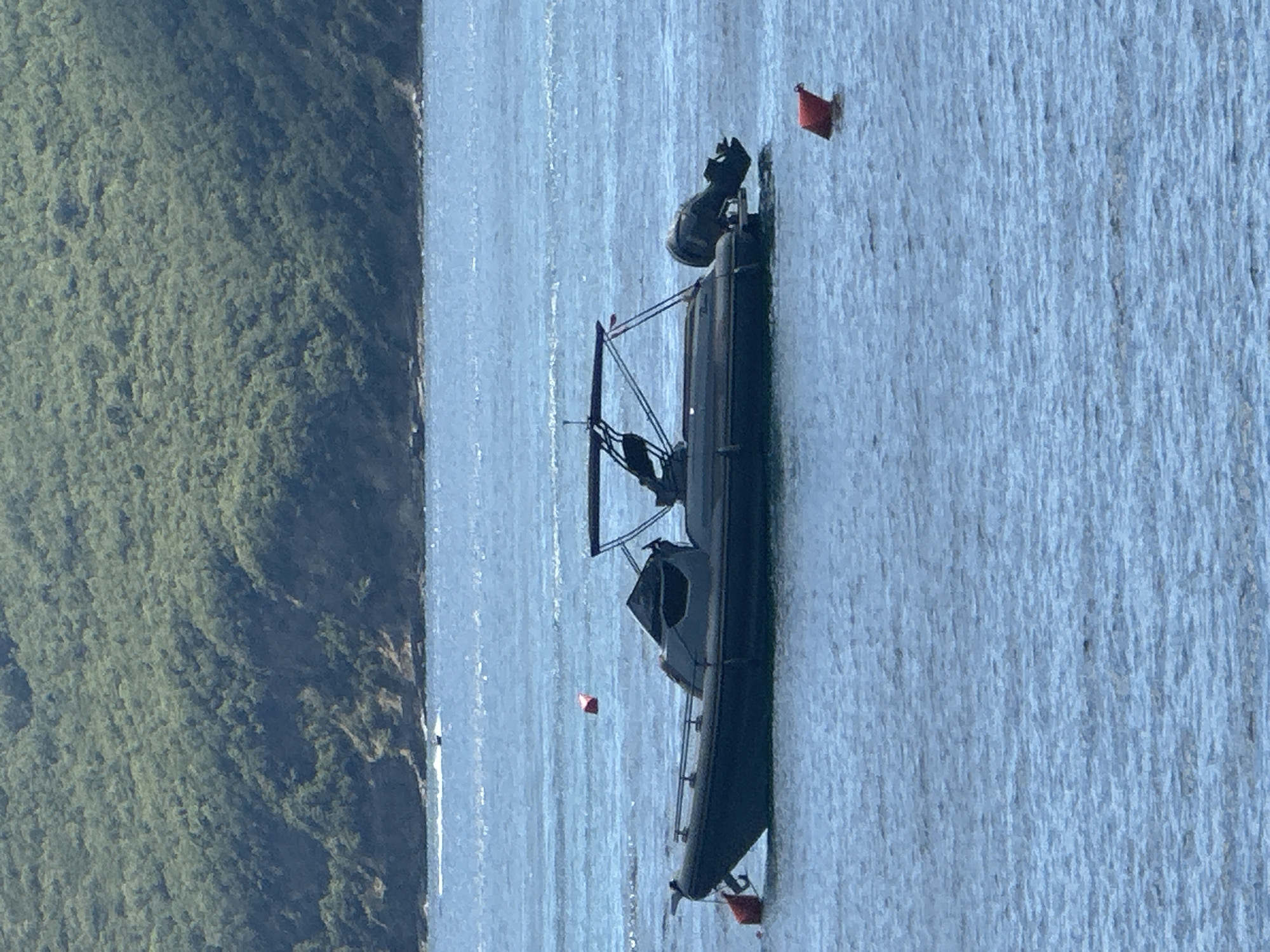 A dark-hulled Rigid Inflatable Boat (RIB) is anchored in calm, blue water, likely off the coast of Budva, Montenegro. The boat has a black canopy supported by metal poles and a visible outboard motor at the stern. Two orange buoys are present in the water: one is near the bow of the boat, and another is further out to the right. In the background, a densely vegetated hillside rises from the water's edge, suggesting a coastal or island landscape. The sky is overcast, casting a diffused light on the scene. No people are visible on the boat or in the water.