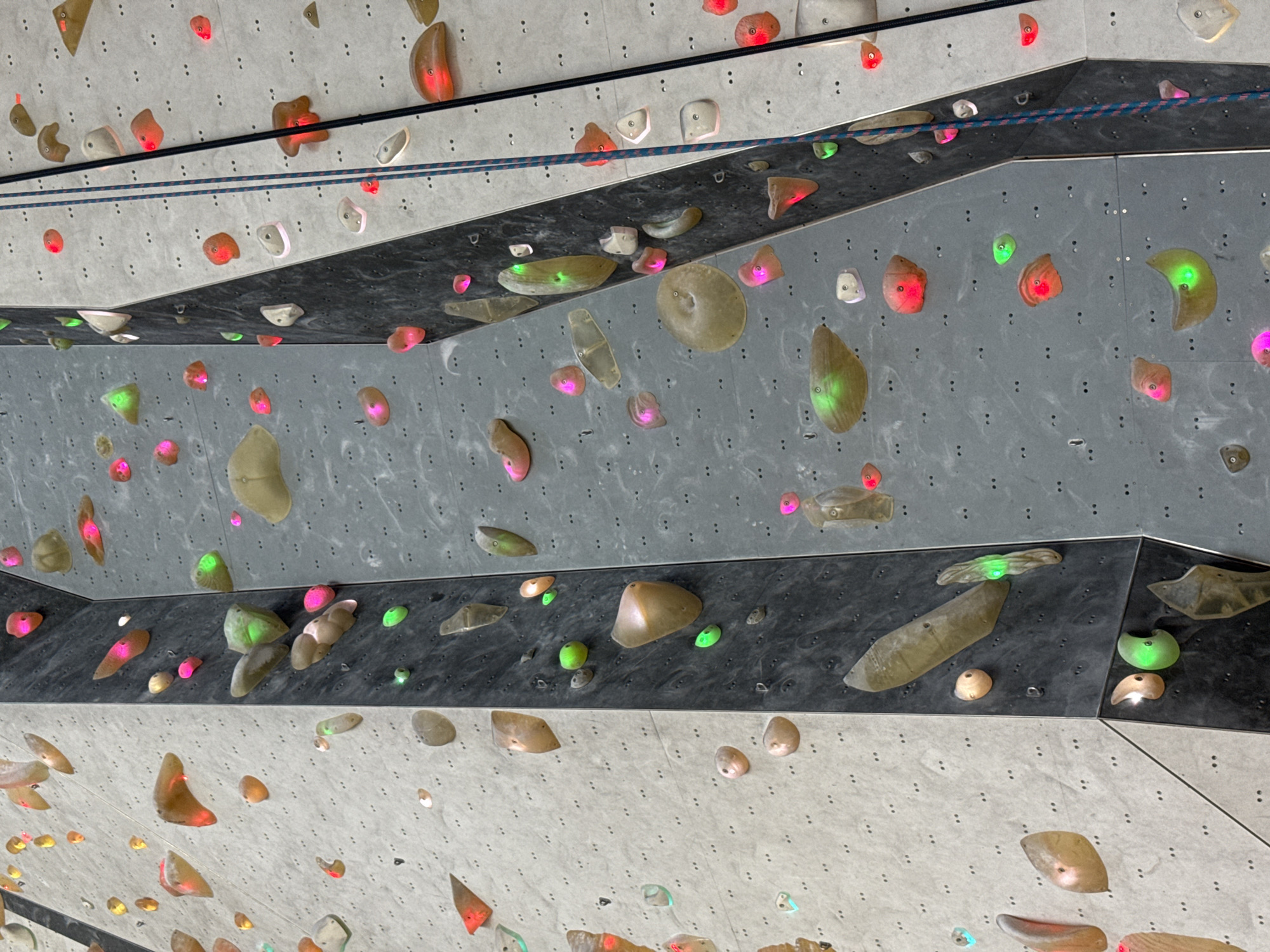 The image presents an upward, close-up view of an indoor climbing or bouldering wall, likely situated in a modern climbing gym in Sofia, Bulgaria. The wall is composed of multiple angular panels in varying shades of gray and black, creating a dynamic, multi-level surface.
Numerous climbing holds of diverse shapes and sizes are affixed to these panels. A distinctive feature is that many holds are translucent and internally illuminated with vibrant LED lights, emitting glowing red, pink, and green hues. This illumination suggests an interactive climbing system, such as a Kilter Board, where specific routes are highlighted by the glowing holds. Other opaque holds in natural tones like brown, beige, and off-white are also interspersed across the wall.
A blue and white striped climbing rope is visible running horizontally across the mid-section of the wall, indicating potential routes or safety lines. Small, regularly spaced attachment points and metal eyelets are visible on the panels.
Although no people are present, the detailed setup of holds and ropes clearly signifies an indoor climbing or bouldering activity. The brightly lit holds create an engaging and technologically advanced atmosphere, characteristic of contemporary climbing facilities. There is no visible text in the image.