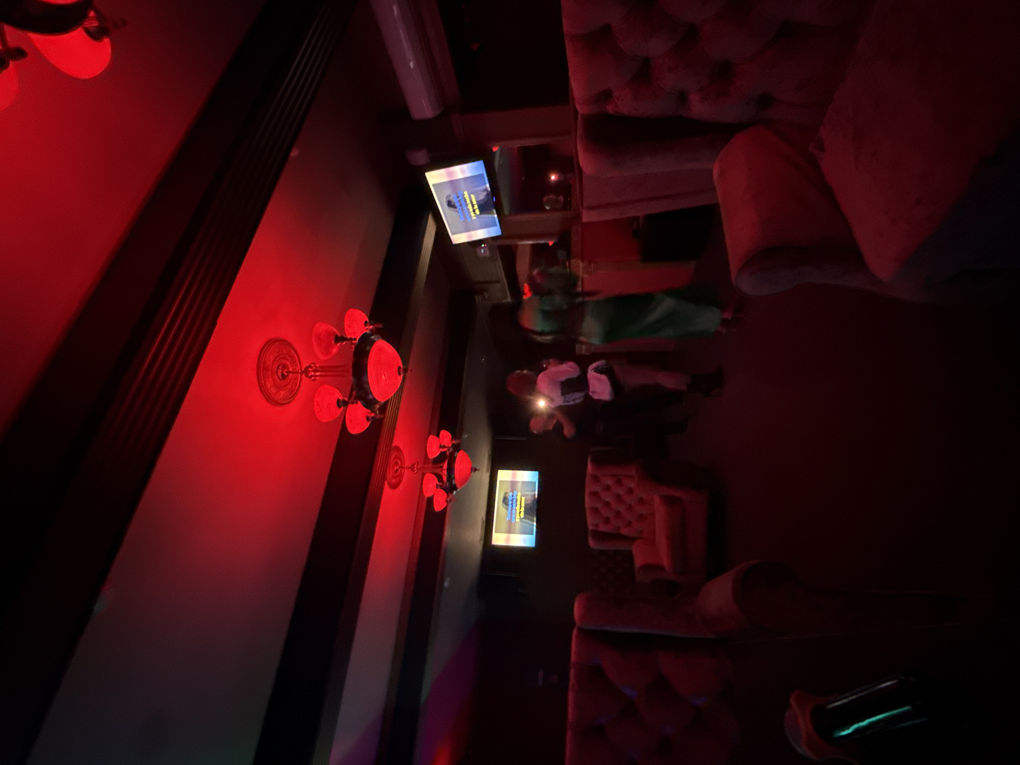 The image captures a dimly lit interior, potentially a lounge or bar, illuminated predominantly by red lighting. Patrons are present, although their activities and interactions are obscured by the low light. Furnishings include upholstered seating and what appears to be a television screen displaying text in a language other than English. The presence of Cyrillic script on the screen and the general architectural style suggest the location is potentially in Moscow, Russia.