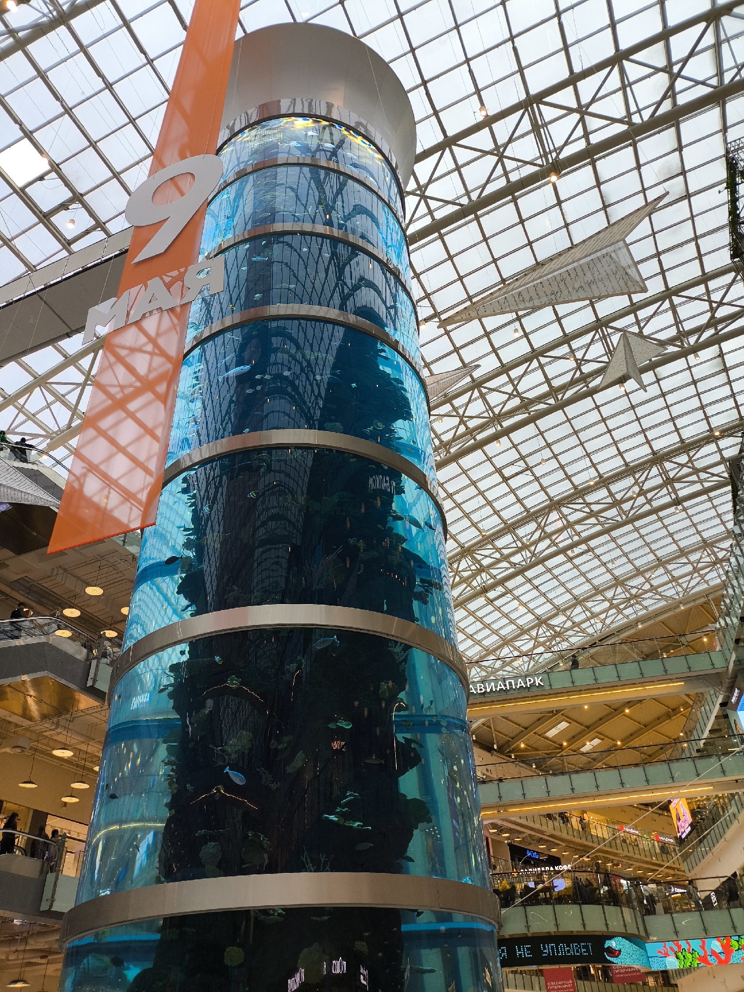 The image depicts the interior of the Aviapark shopping mall in Krasnogorsk, Russia. The focal point is a large, cylindrical aquarium extending upwards towards the mall's glass ceiling. The aquarium contains fish and aquatic plants. An orange banner with the number "9" and the Russian word "MAЯ" (May) hangs adjacent to the aquarium. Above, the mall's glass roof is supported by a network of metal beams. Decorative paper airplanes hang from the ceiling. In the background, multiple levels of the mall are visible, with lights and signage, and people can be seen on the various levels.