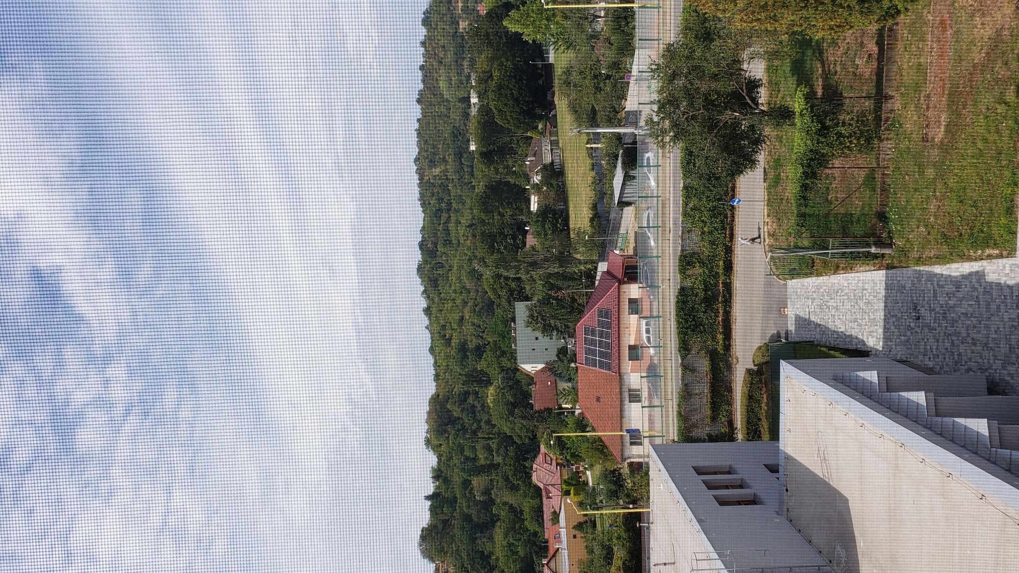 The image, taken from a high vantage point through a window screen, depicts a suburban landscape. In the foreground, the roof and side of a modern, grey building are visible, with a patterned concrete walkway leading away from it. To the right of the walkway is a grassy area with a fence and some trees.
Further into the scene, a paved road runs horizontally, with several parked cars visible on the other side. Beyond the road, a residential area is characterized by houses with red tiled roofs, some featuring solar panels. A larger, light-colored building with multiple windows is also present among the houses.
The middle ground is dominated by a lush, green, tree-covered hillside. The background consists of a sky with scattered white clouds. A lone figure is walking on the paved road in the middle distance. The overall impression is of a calm, daytime scene in a suburban environment, likely Prešov, Slovakia, given the architectural style and vegetation.