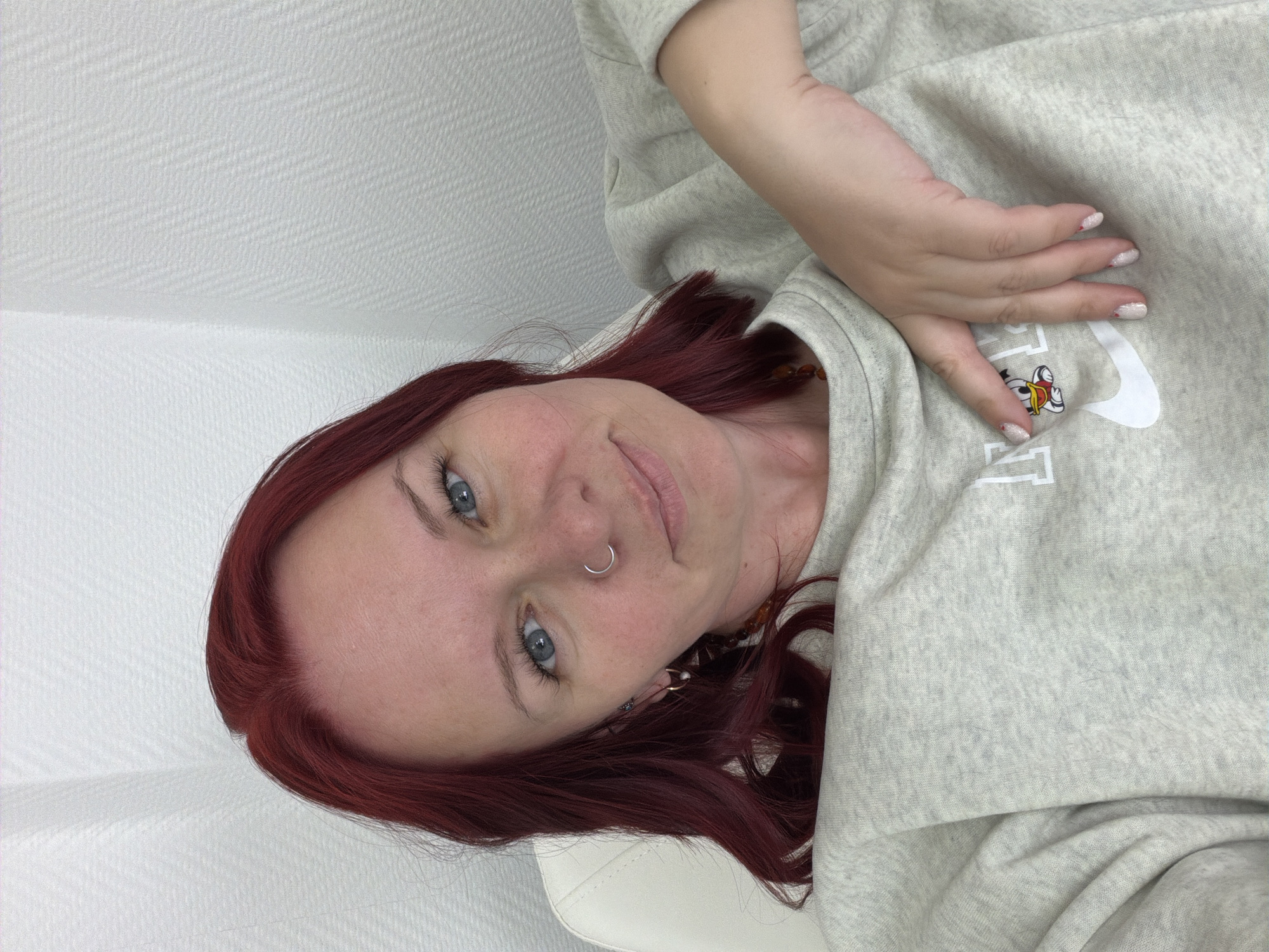 An indoor scene in Chaikovsky, Russia, depicts an adult female positioned off-center, leaning against a light-colored, possibly upholstered, chair or surface. She has shoulder-length, dark red hair and blue eyes, and a silver-toned hoop nose ring is visible on her right nostril. Small hoop earrings are present, with a pearl-like drop on her left ear. Her fingernails are manicured with light-colored polish and small red decorative elements. She wears a light grey, crew-neck sweatshirt featuring a white "NIKE" logo on the left chest, below which a small cartoon character resembling Donald Duck is depicted. A necklace composed of amber-colored, irregularly shaped beads is visible around her neck. Her left hand rests casually on her chest, near the sweatshirt's logo. The background consists of a uniformly white, textured wall, illuminated by soft, ambient lighting. The subject is looking directly at the viewer with a slight, closed-lip smile.