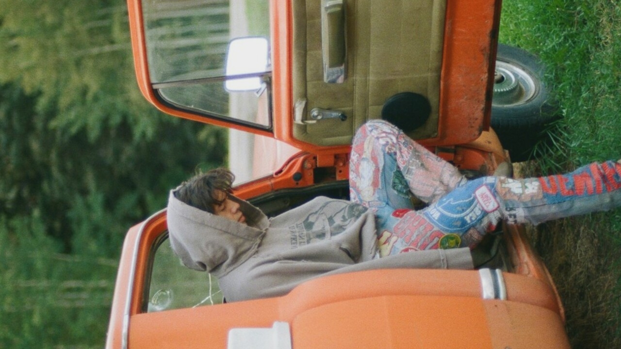 An individual wearing a grey hooded sweatshirt and colorful, patchwork pants sits in the open driver's side door of an orange pickup truck. The individual's head is bowed, and their face is not clearly visible. The truck is parked on a grassy area, with trees in the background. The scene appears to be outdoors, possibly in a rural or park-like setting. There is no indication of a specific location within Moscow, Russia, or any other city.