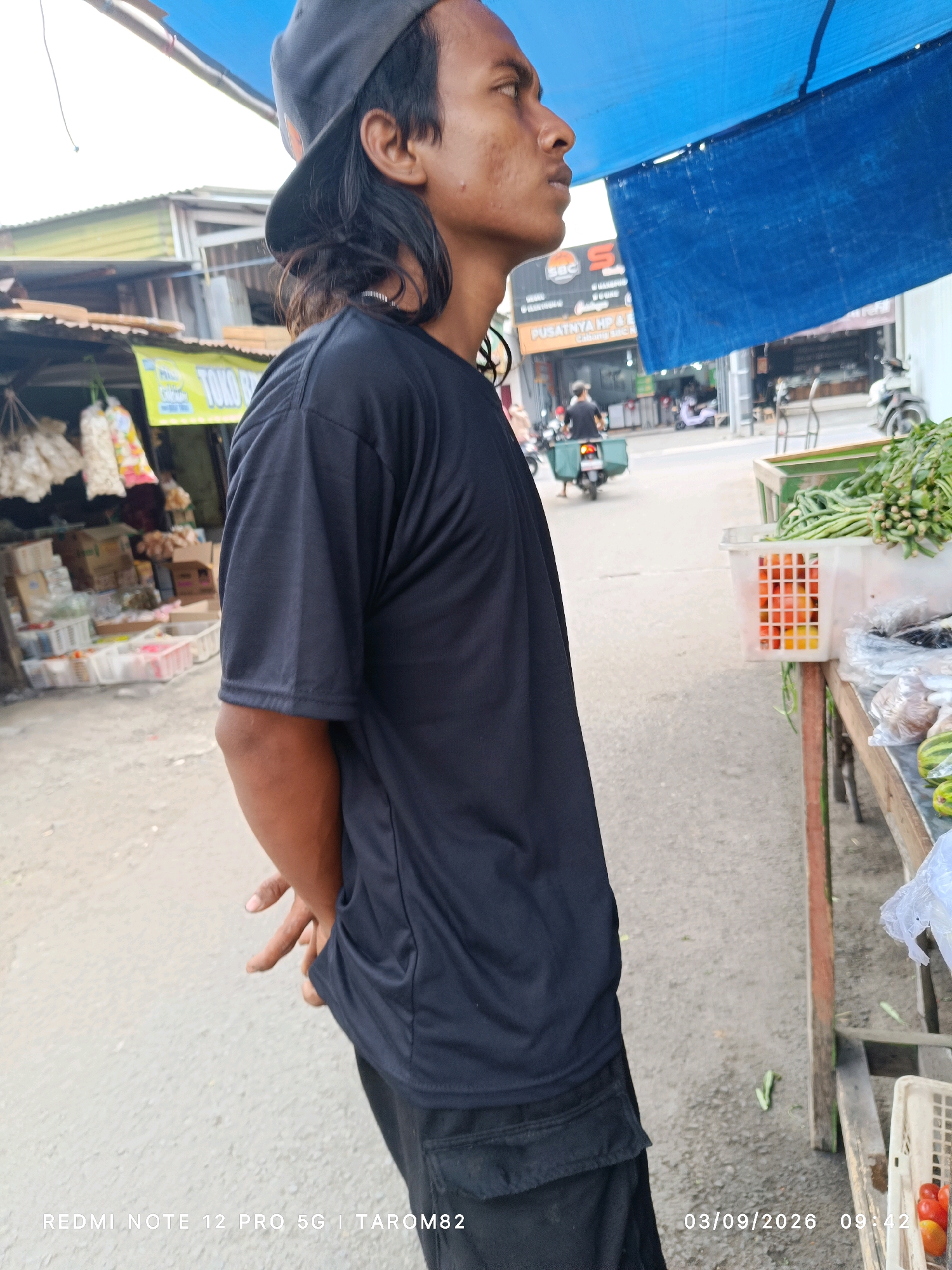 The image depicts a street scene in Kandangan, Indonesia, likely a market area. A young man wearing a black t-shirt and a dark baseball cap is the primary subject, positioned in the foreground, facing right and looking away from the camera. His hands are clasped behind his back. Behind him, the street is lined with small shops and market stalls displaying various goods, including vegetables and packaged items. A blue tarp is stretched overhead, providing shade. In the background, a motorcycle with a passenger is visible, suggesting general street activity. The ground is paved with asphalt, and the overall environment appears to be an outdoor commercial space.