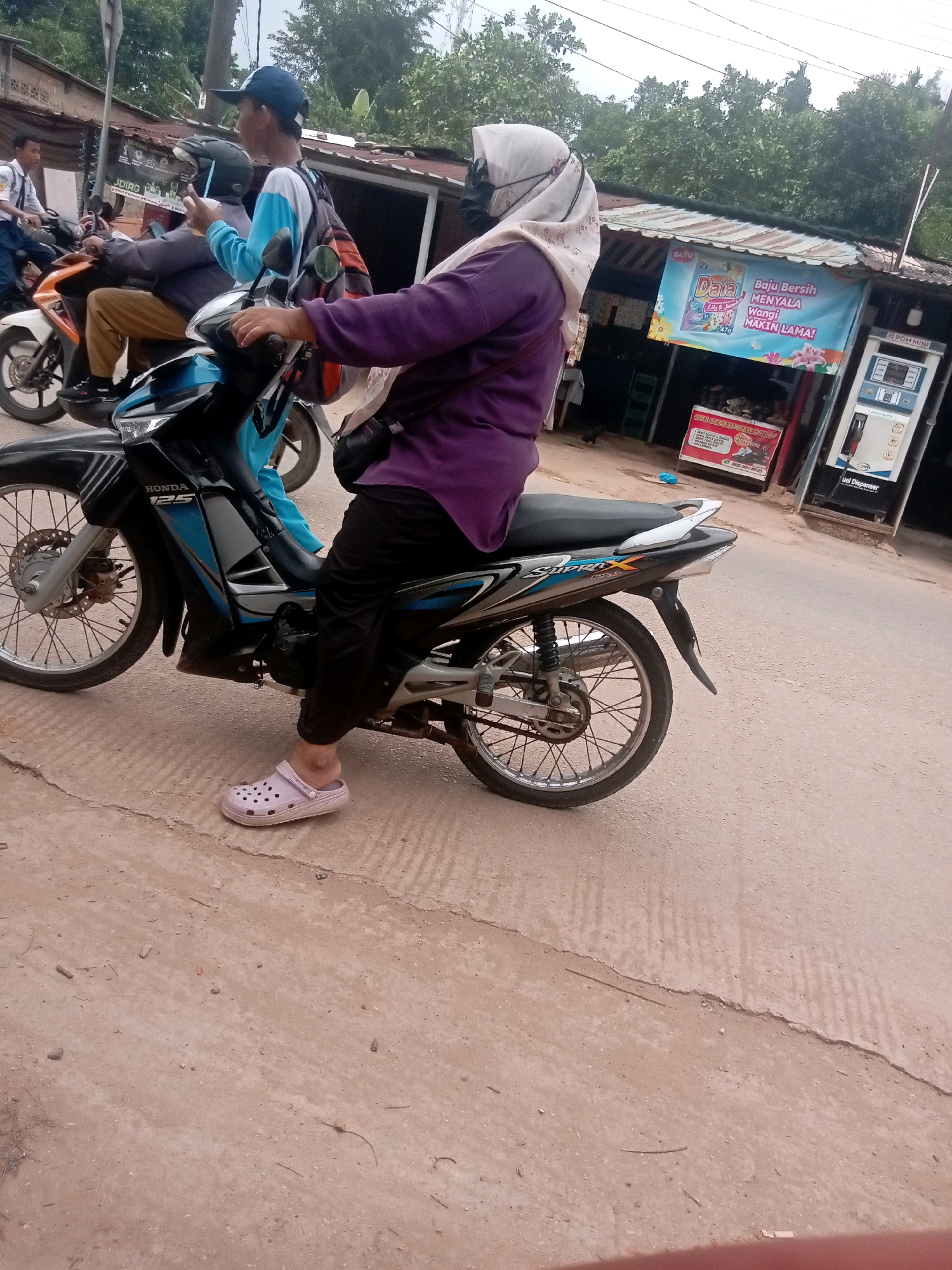 The image shows a street scene in Batam, Indonesia, with several people on motorcycles. In the foreground, a woman wearing a purple top, black pants, and a light-colored hijab is riding a black Honda motorcycle. She is wearing light pink Croc-style shoes. To her left and slightly behind her, another person on a white and orange motorcycle is visible, wearing a blue and white shirt and a backpack. Further in the background, a person in a school uniform is riding a motorcycle. Behind the motorcycles, there is a roadside store with a sign advertising laundry detergent. A fuel dispenser is also visible to the right of the store. The ground is paved with asphalt and concrete, showing signs of wear and tear, with some cracks and debris. The overall environment suggests a typical street scene in a town or city in Indonesia, with a mix of daily activities and commercial establishments.