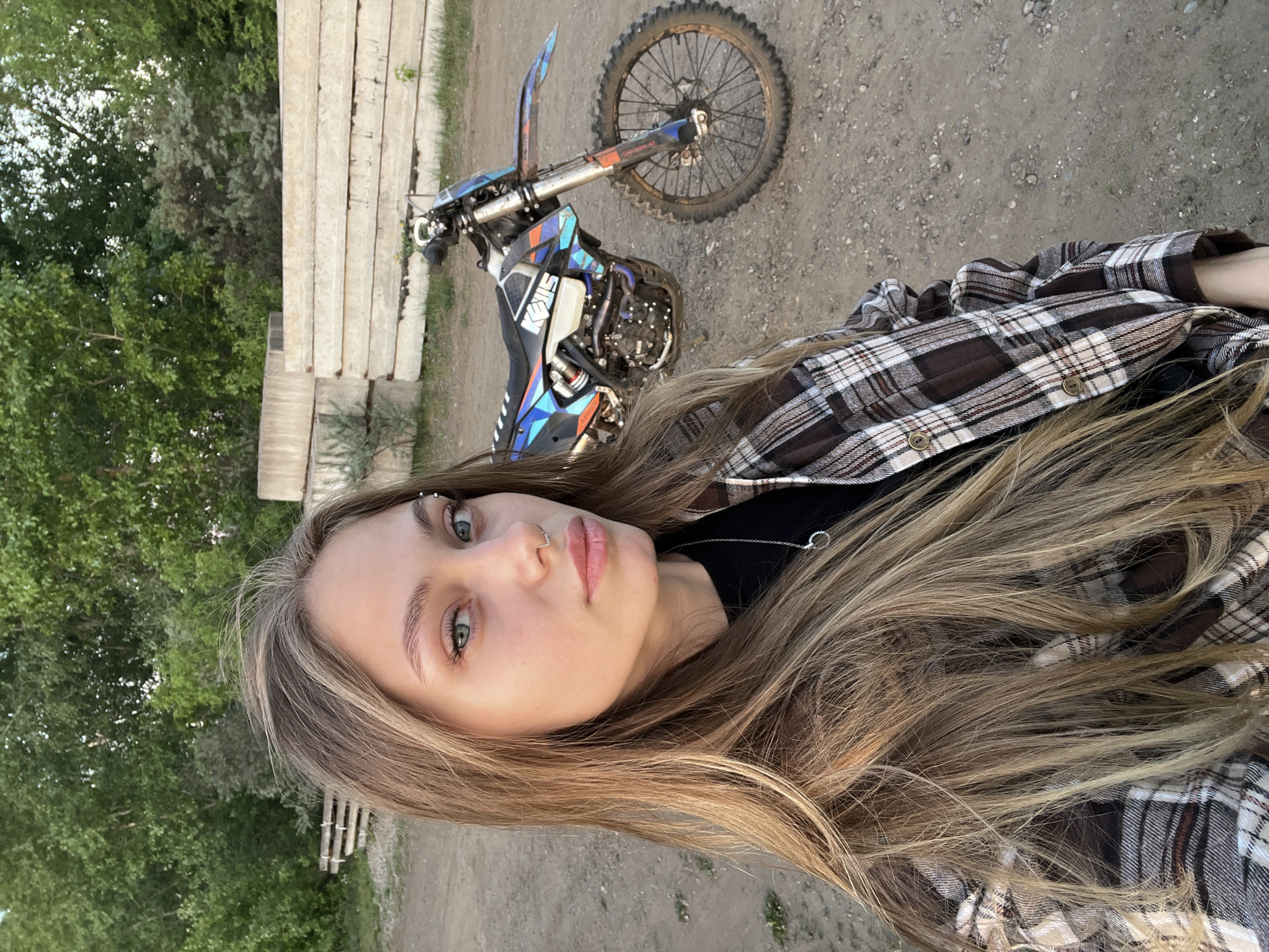 A woman poses in front of a dirt bike, likely outdoors. She has long blonde hair, and wears a nose ring and an eyebrow piercing. The dirt bike is blue and orange. The background shows a dirt surface, trees, and concrete blocks. There is no explicit information in the image to confirm that this is Chaikovsky, Russia.