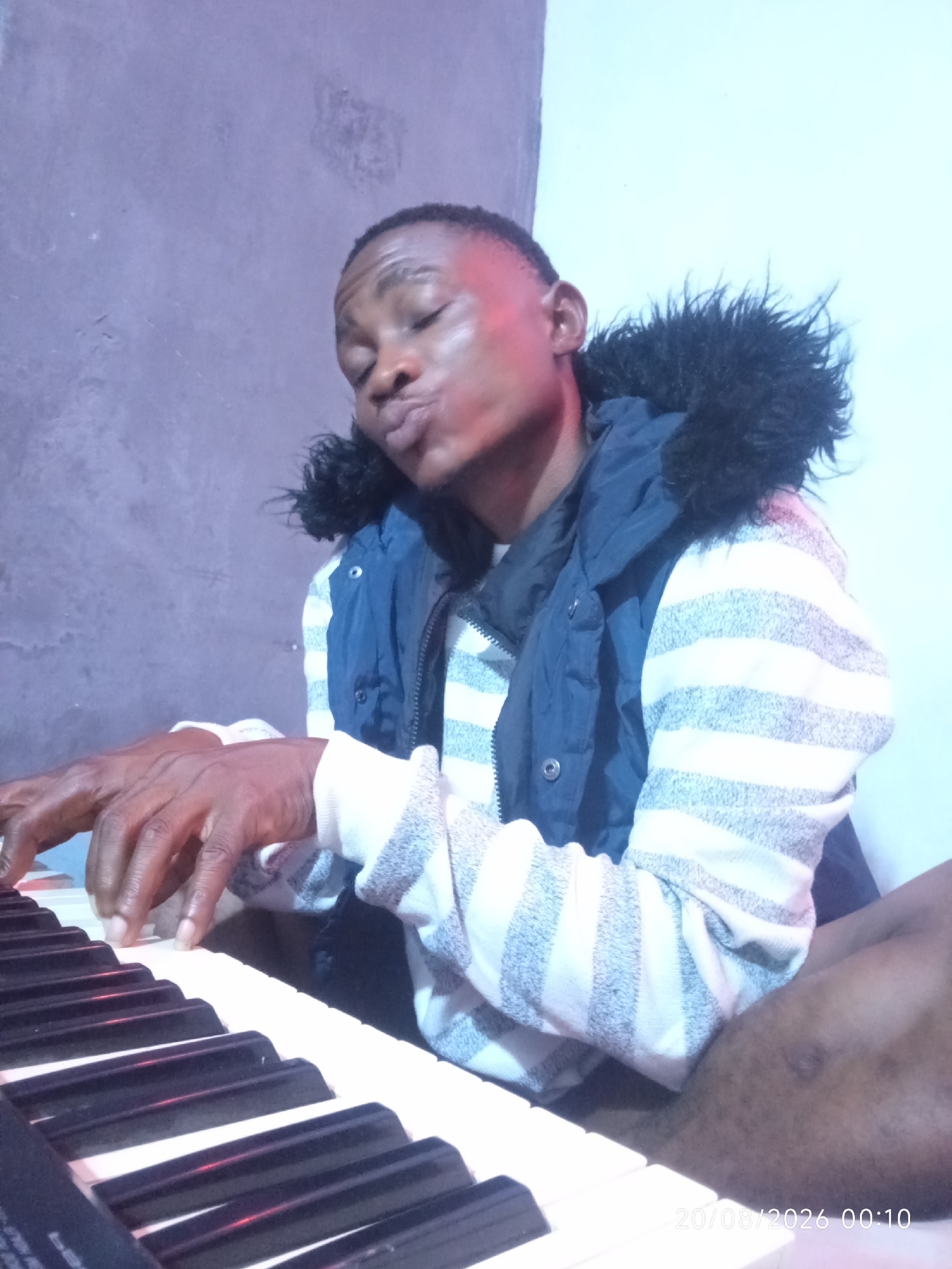 A dark-skinned man with short hair is sitting in front of a keyboard, playing it with his hands. He is wearing a blue vest over a striped long-sleeved shirt with a fur-lined hood. The man has his eyes closed and his lips pursed as if he is singing or deeply concentrating on the music. He is positioned to the left of the frame, with the keyboard taking up the lower left portion. The background is a plain, textured wall, predominantly purple with some lighter blue and white areas. A timestamp in the lower right corner reads "20/08/2026 00:10". The scene appears to be indoors, possibly in a bedroom or a studio in Adankolo, Nigeria.