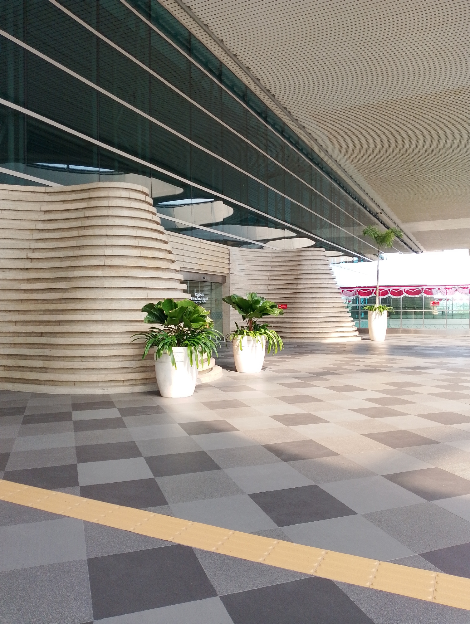 The image captures the exterior of Yogyakarta International Airport in Palihan, Indonesia. The building features a modern architectural style with a curved, layered facade made of light-colored stone or concrete. Large glass windows run along the upper portion of the building, reflecting the sky. In front of the building, several large white planters hold lush green plants, contributing to the landscaping. The ground is paved with a checkered pattern of light and dark gray tiles, with a tactile paving strip in yellow running diagonally across the foreground. A few potted palm trees are also visible, adding to the tropical ambiance. In the background, a red and white striped canopy suggests an outdoor waiting area or event space. The lighting indicates it is daytime, with sunlight casting shadows on the pavement. There are no people visible in the scene.