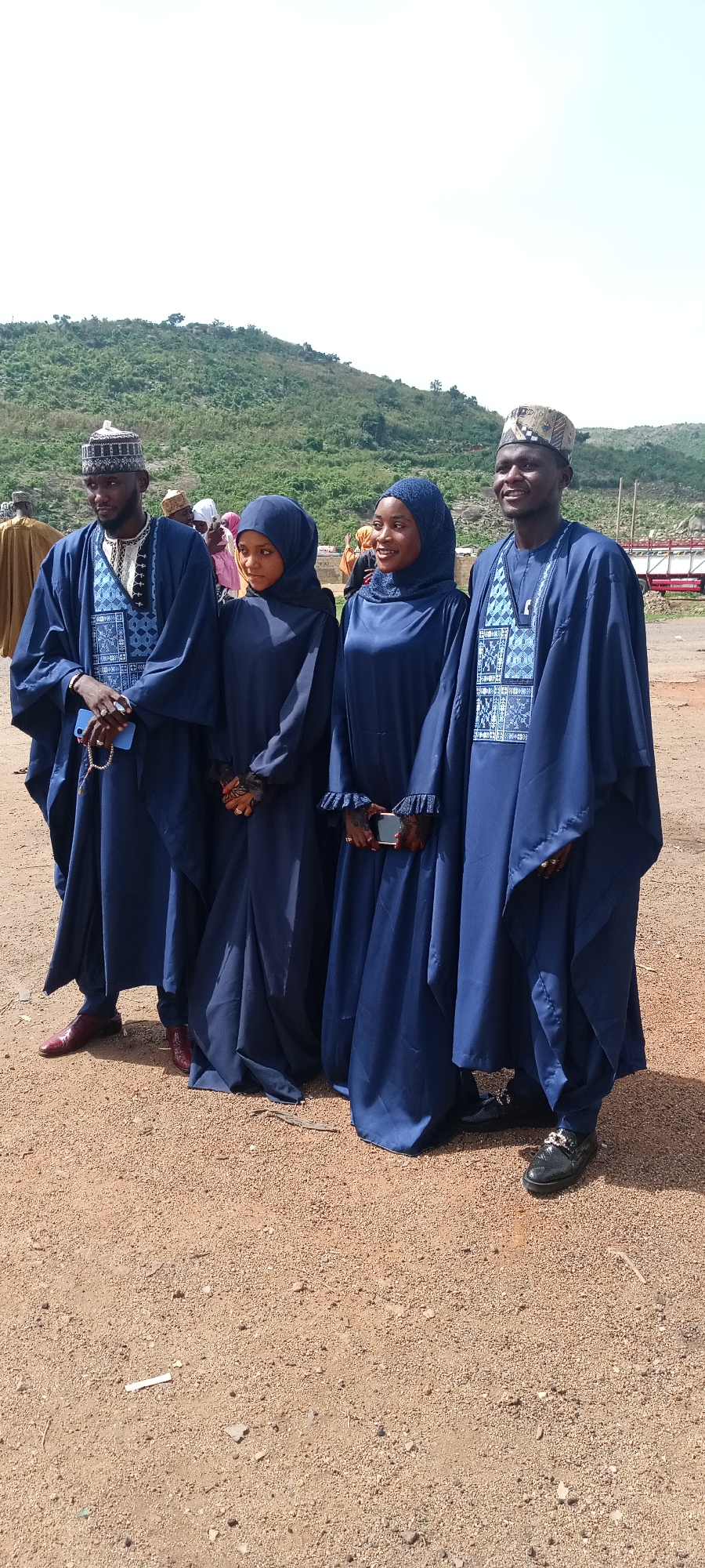 Four people are standing outdoors in Naraguta Hausawa, Nigeria. Two men and two women are dressed in blue, traditional Hausa clothing. The men wear embroidered caps and robes with patterned vests. The women wear hijabs and long dresses, also in blue. The ground is sandy. In the background, there is a hill covered in vegetation and a glimpse of a red and white structure.