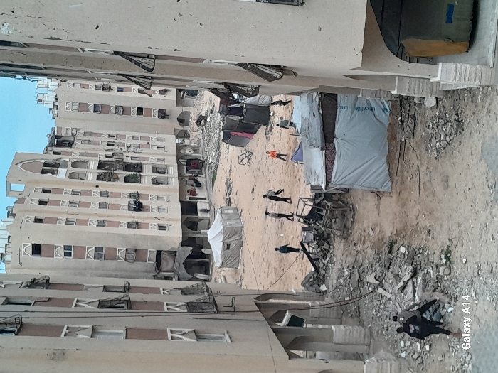 The image depicts a devastated urban residential area in Khan Yunis, Palestinian Territory, characterized by extensive structural damage and makeshift living conditions. Multiple multi-story apartment buildings, varying in height and design, line a wide dirt-covered courtyard or open space. These buildings show severe damage, including numerous shattered windows, pockmarked facades indicative of bullet impacts or shrapnel, and visible interior destruction. Some balconies are damaged, and window shutters hang loosely or are missing.
On the ground, piles of concrete rubble, twisted metal, and general debris are scattered throughout, particularly in the foreground and near the base of the damaged structures. Several temporary shelters are erected in the open space. A large, light-colored canvas tent stands prominently in the middle-left. Near the right, a smaller, makeshift shelter is constructed from tarpaulins, with a blue tarp displaying the "unicef" logo. Clothes are visible hanging on lines near some of these shelters.
Approximately ten individuals, appearing to be mostly children or young adults, are present in the scene. A group of six individuals walks across the dirt area in the middle ground, one wearing an orange shirt, the others in dark clothing. In the lower foreground, two people in dark clothing stand near a pile of rubble. The ground consists of uneven, dry dirt with scattered stones and debris. The sky above is overcast, providing diffuse lighting to the scene.