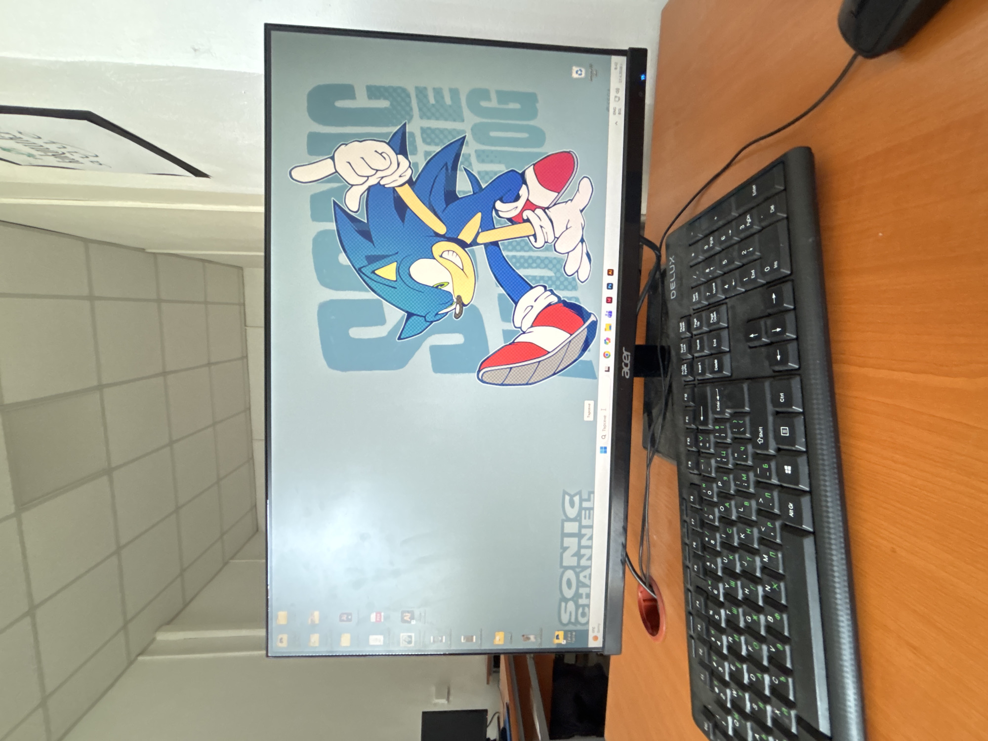 The image captures a computer workstation set up in an indoor environment, likely in Troyan, Bulgaria, as indicated by the Cyrillic characters on the keyboard and the Bulgarian word "Търсене" (Search) on the monitor.
The central feature is a black Acer monitor, turned on and displaying a colorful wallpaper of Sonic the Hedgehog. Sonic is depicted in a dynamic pose, suspended in mid-air, with his signature blue quills and red and white shoes. The wallpaper's background features the text "SONIC CHANNEL" vertically on the right and a mirrored "SONIC" horizontally at the top. The Windows taskbar is visible at the bottom of the screen, showing several application icons and a search bar with "Q Търсене".
To the right of the monitor, a black DELUX keyboard rests on a light brown wooden desk. The keyboard clearly displays both Latin and Cyrillic alphabets on its keys. Further to the right, a black wired computer mouse is visible. Wires for the monitor, keyboard, and mouse are present on the desk, with a red circular cable management grommet located to the left of the keyboard.
The desk itself is made of light-colored wood. In the background, a light-colored wall with a grid-like pattern, possibly tiles or panels, extends upwards and to the left, where it appears to transition into a stepped or angled architectural feature. The overall lighting suggests an indoor setting, likely during the daytime. No human activity is depicted; the focus is solely on the workstation setup.