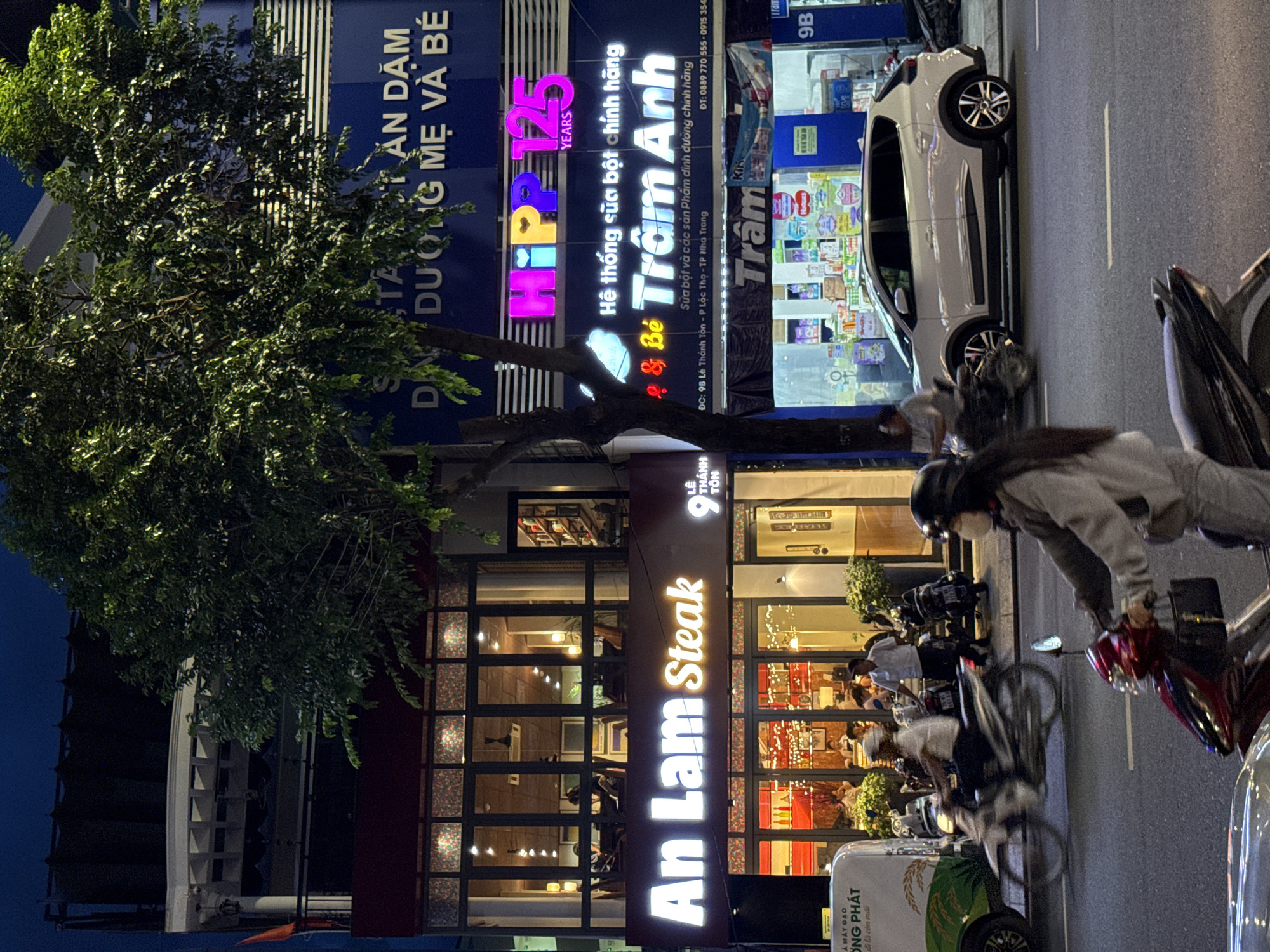 This is a nighttime street-level photograph taken in Nha Trang, Vietnam, featuring several commercial establishments and traffic. The foreground shows a street with two people on scooters. The person on the closer, red scooter is wearing a helmet and a grey jacket, with a black bag hanging from the handlebars. They are in motion, moving towards the left of the frame. Behind them, a person on a white scooter, also wearing a helmet, is also in motion.
The background is dominated by the facades of three buildings. The leftmost building is a restaurant called "An Lam Steak," indicated by a prominent illuminated sign. Its upper floor has large windows revealing patrons seated inside. The middle building has a bright, multi-colored neon sign reading "HIPP 125 YEARS" and beneath it, signage for "Trầm Anh," which appears to be a children's nutrition store, with shelves of products visible through its window. The rightmost building is partially obscured by a tree but displays a blue facade with white horizontal stripes and some signage.
The street is paved and appears to be well-lit by the signs and interior lights of the businesses. A white delivery van is parked on the left side of the street, near the "An Lam Steak" restaurant. The overall atmosphere is urban and active, typical of a commercial street in the evening.