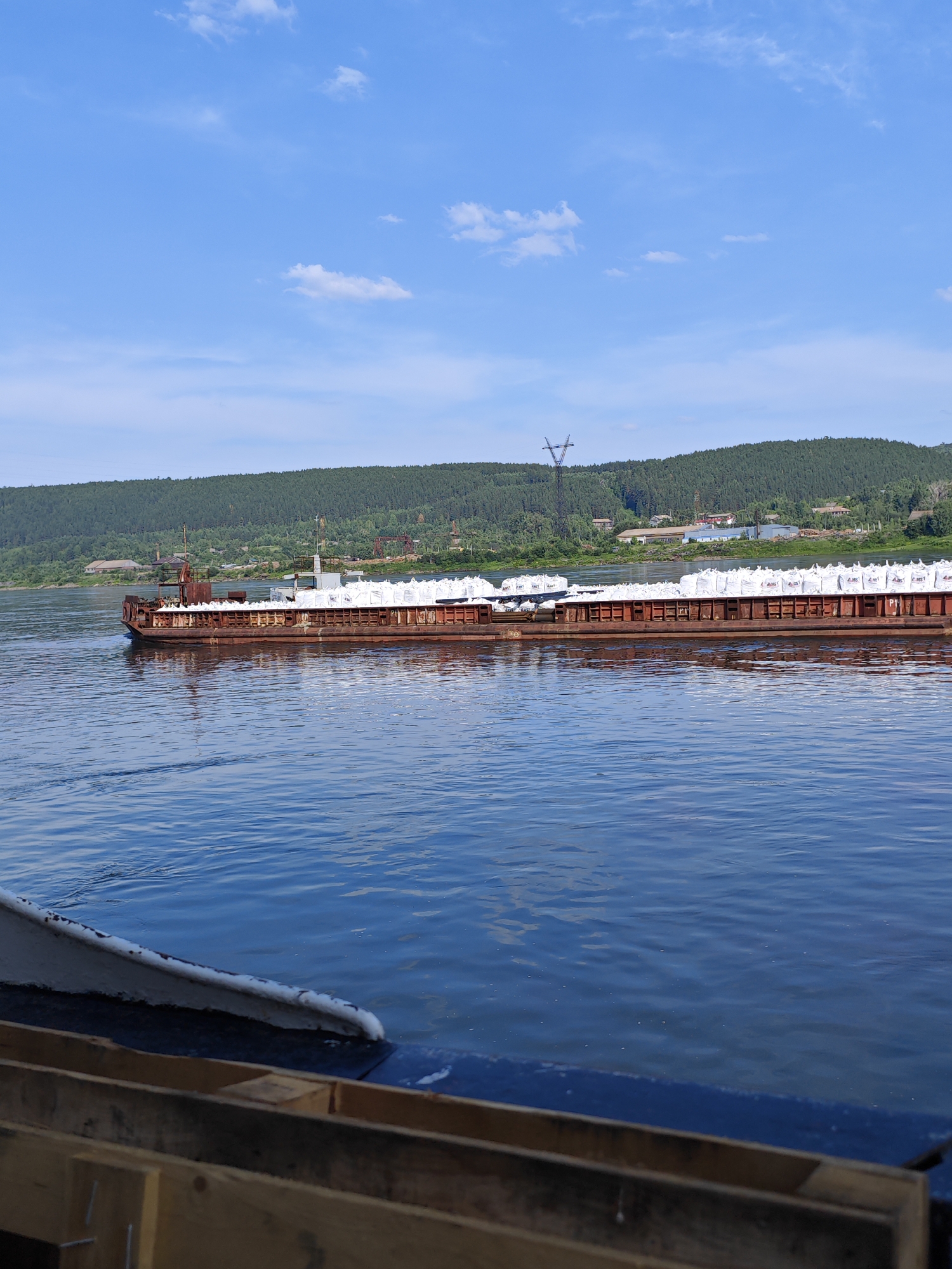 An outdoor, low-angle, medium shot captures a barge laden with white sacks on a wide, blue river. The barge, primarily a rusty red color with some white accents, is moving from left to right in the frame. The water's surface exhibits gentle ripples, reflecting the blue sky and scattered white clouds above. In the background, a lush green, tree-covered hill slopes down towards the river, dotted with small buildings and industrial structures, including a tall transmission tower. The foreground features the edge of another vessel, with visible wooden crates and a dark blue hull. The scene appears to be set in the city of Pristan', Russia, given the context of the barge traffic on the river and the surrounding landscape. There are no people visible in the image.