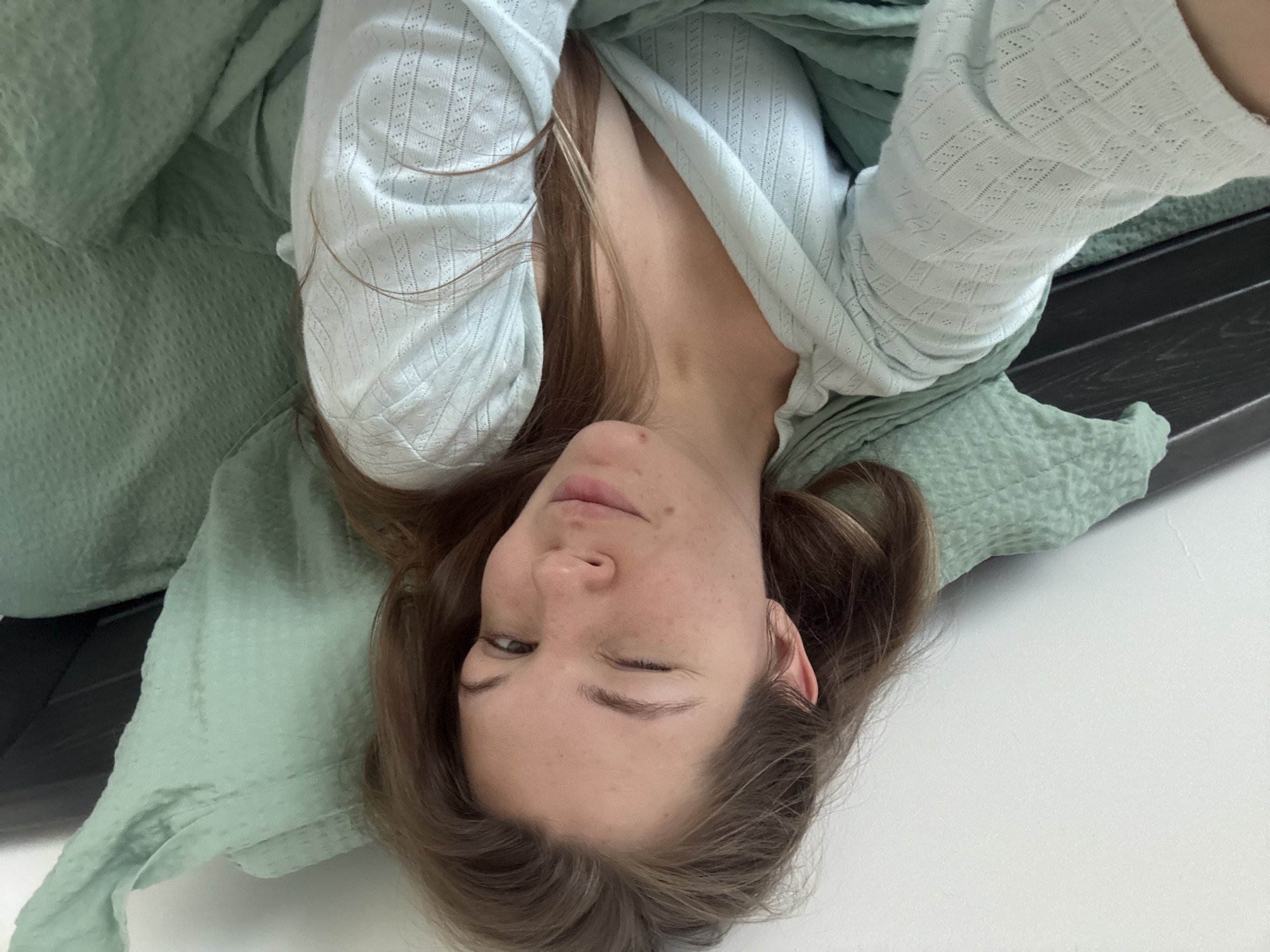 This image depicts a young adult female lying in a bed within the city of Vozdovac Urban Municipality, Serbia. The subject, with long, light brown hair, is positioned for a selfie, looking directly at the camera. Her left eye (viewer's right) is closed in a wink, while her right eye is open. Her lips are slightly pursed. She is wearing a pale greenish-blue, long-sleeved top featuring a textured, open-knit diamond pattern and a V-neckline that reveals her upper chest. The bedding consists of a mint green quilted duvet or blanket and a pillow of the same color and texture. A dark brown or black wooden headboard is partially visible behind her on the right side of the frame, and the background wall is white. The scene is illuminated by soft, diffuse light.
