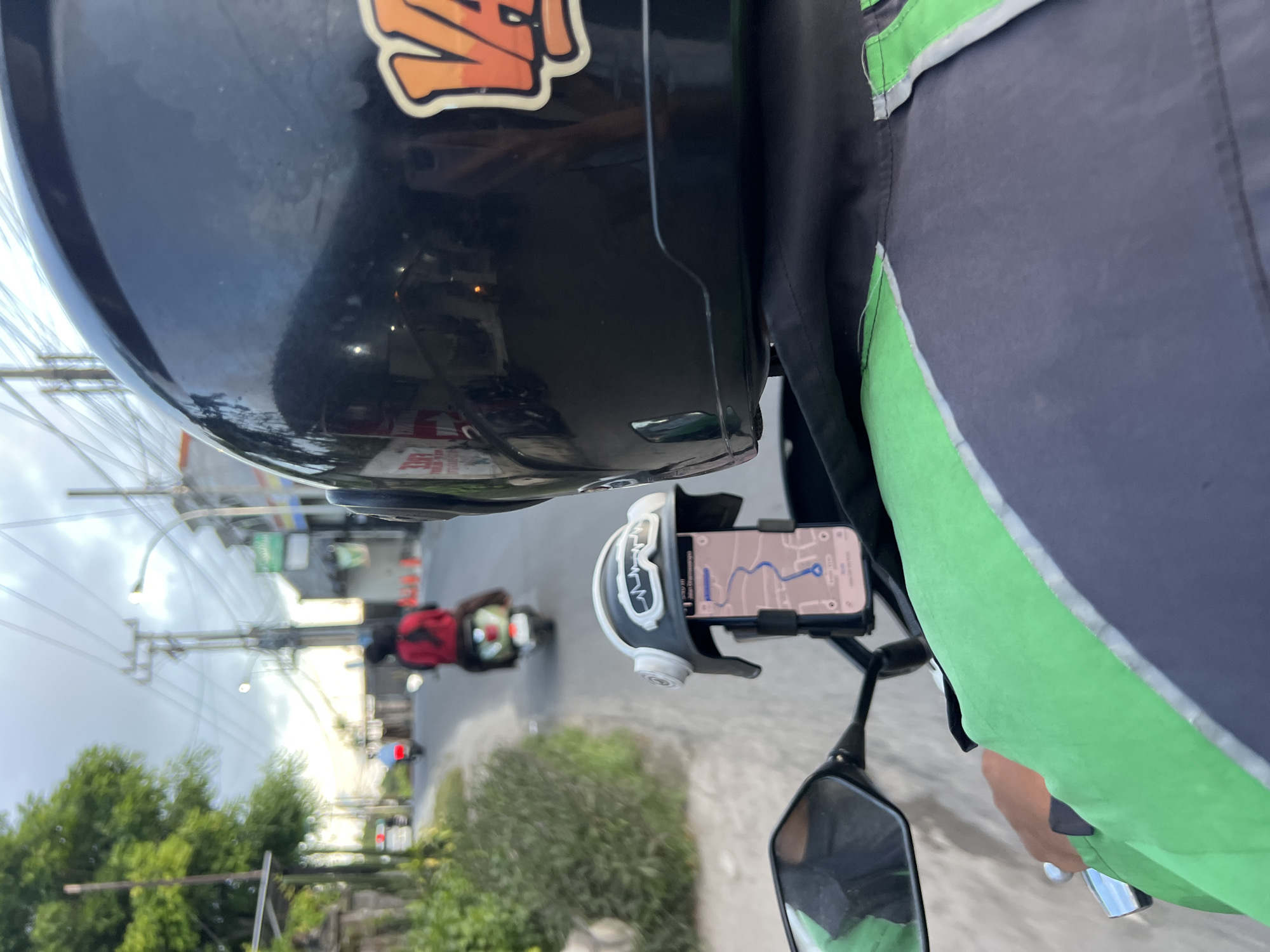 A street-level, point-of-view shot from a motorcycle, shows a rider wearing a black helmet with an orange graphic on the top and a green and black jacket, navigating a city street in Unknown, Unknown. A phone mounted on the motorcycle's handlebar displays a map with a blue route. In the background, another motorcycle with a rider wearing a red jacket is visible further down the road. The environment includes buildings, trees, and streetlights.