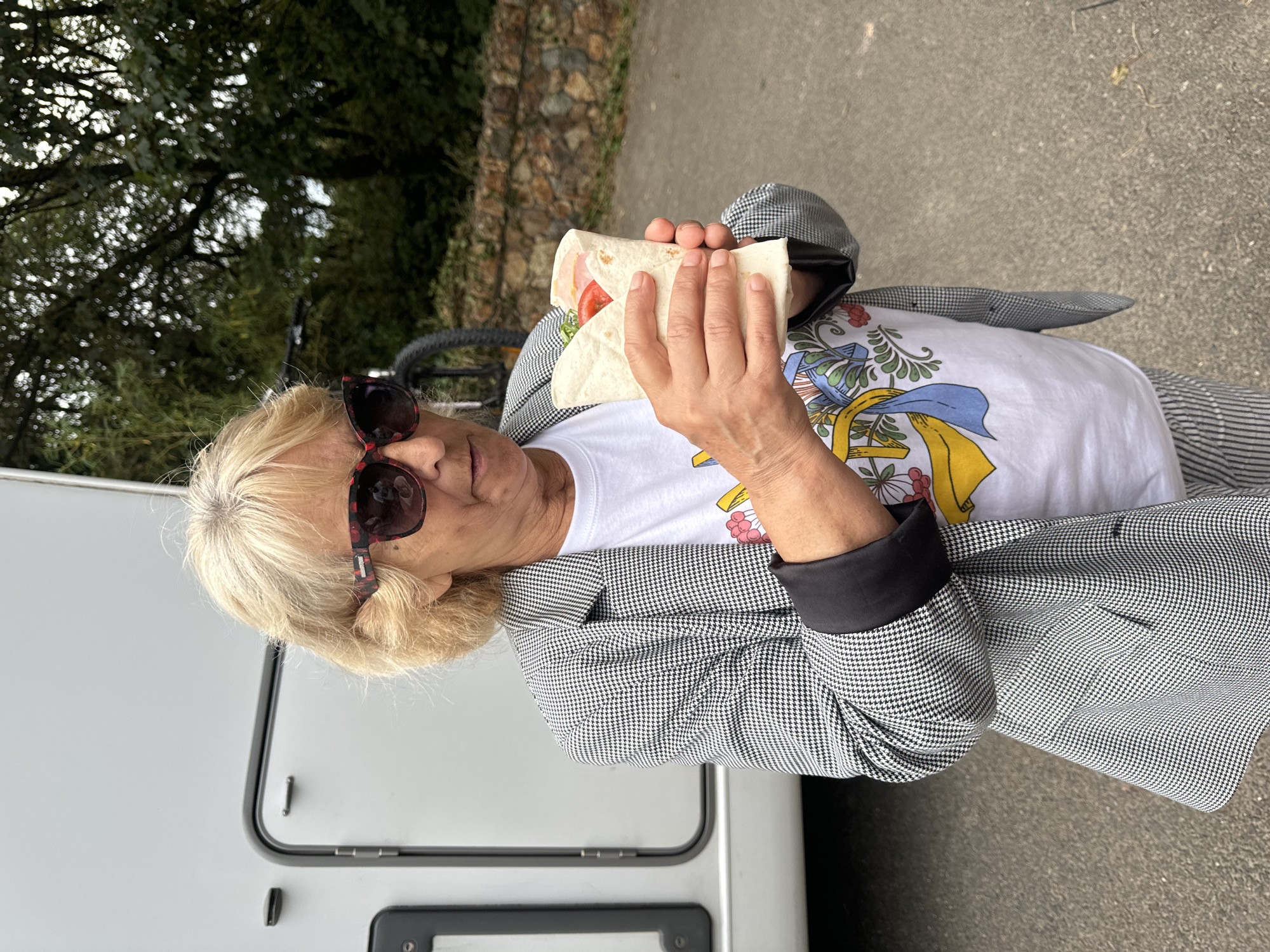A middle-aged woman with blonde hair, wearing sunglasses with red and black frames, a black and white houndstooth blazer over a white t-shirt with a colorful graphic print, and striped pants, stands outside holding a wrap sandwich. She is positioned in front of a white RV, with a bicycle visible behind her to the left. The background consists of lush green trees and a stone wall. The setting appears to be a park or campsite, possibly in Courtown, Ireland, given the natural, outdoor environment.