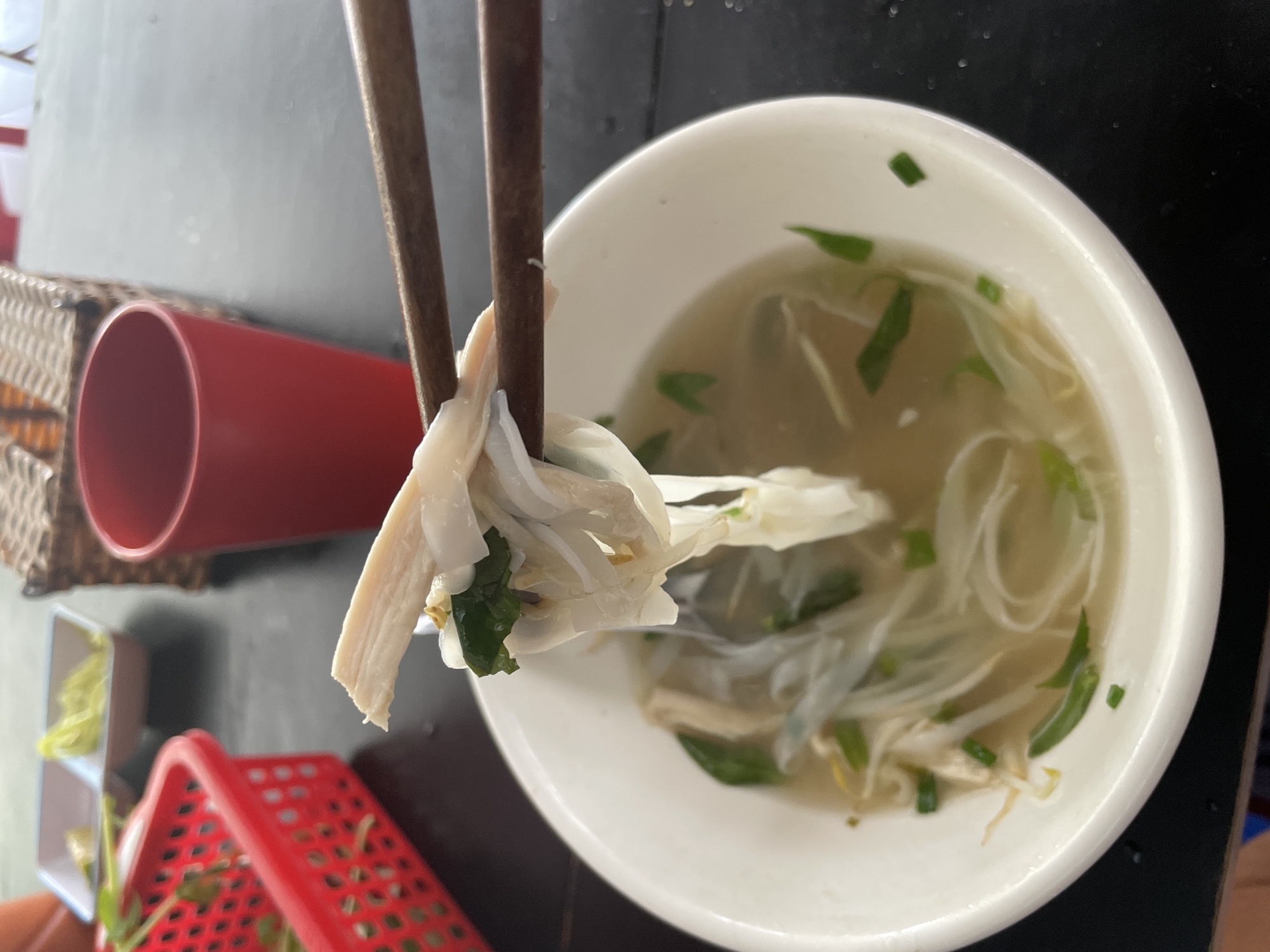 In Phu Thuy Ward, Vietnam, a close-up shot captures a bowl of pho being lifted by chopsticks. The pho contains rice noodles, thinly sliced chicken, bean sprouts, and green onions in a clear broth. In the background, a red plastic basket with some green garnish sits on a dark table next to a woven basket and a red cup. The scene appears to be set in an outdoor dining area, as suggested by the natural lighting and the presence of other diners' belongings.