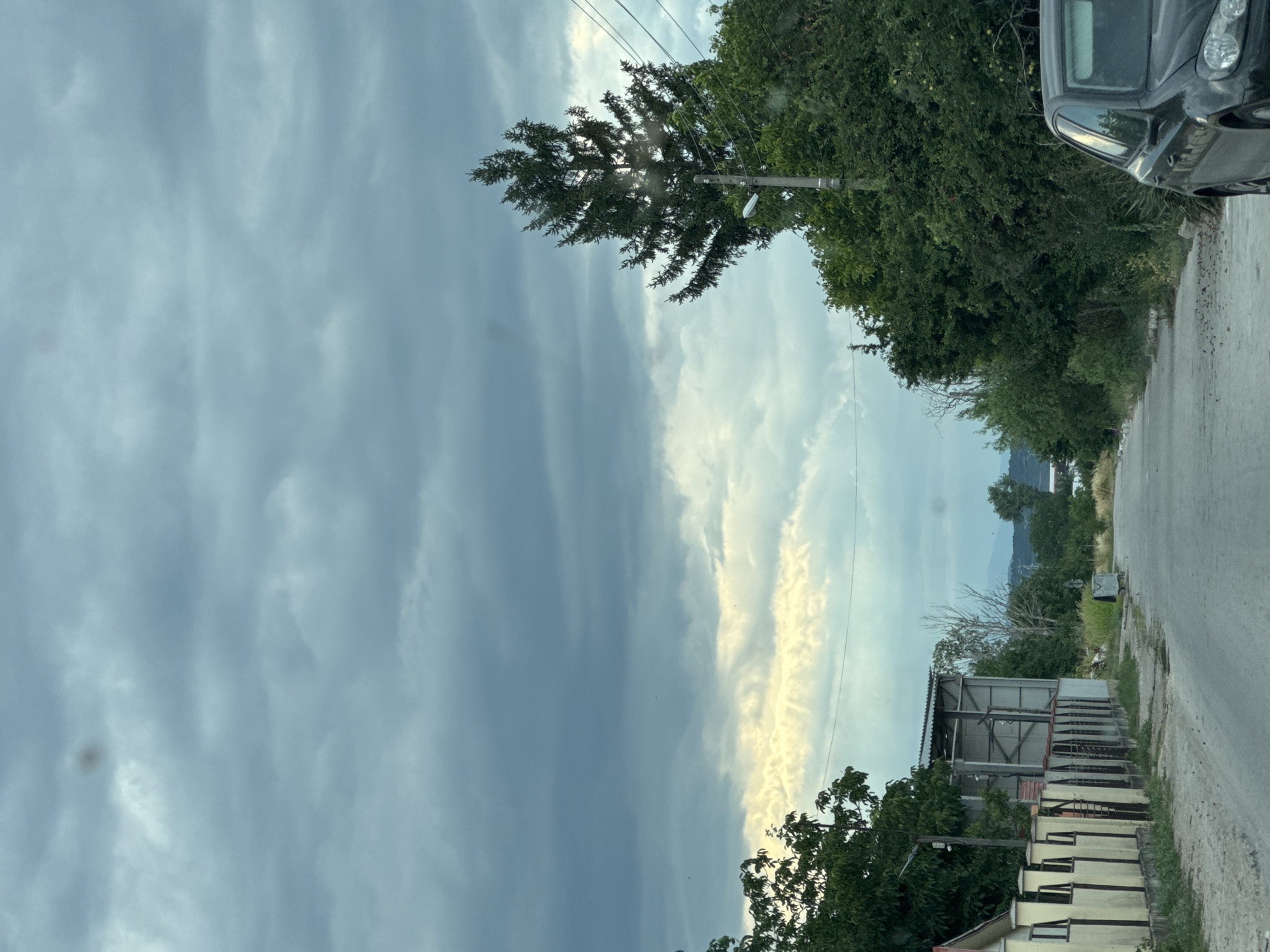 This is a vertical, medium-shot photograph taken from the perspective of someone in a car, looking out at a rural roadside scene. The sky dominates the left side of the frame, filled with dramatic, layered grey clouds interspersed with a bright, yellowish-white band of clouds, suggesting either sunrise or sunset with varying cloud cover.
On the right side of the image, a paved road runs diagonally into the distance. Along the side of the road, there is a dense line of green trees and vegetation. To the right of the road, a dark-colored car is partially visible in the upper right corner. Further down the road, there are several structures, including a building with a corrugated metal roof and a light-colored wall with barred windows, and a smaller grey bin. A utility pole with a light fixture stands near the trees. In the far distance, a hazy outline of mountains can be seen against the sky.
The overall impression is a slightly overcast day, possibly late afternoon or early morning, in a rural area. The sun's rays are breaking through the clouds in one section, creating a bright contrast. There is no visible text in the image. The scene is quiet and appears to be a view from a moving vehicle.