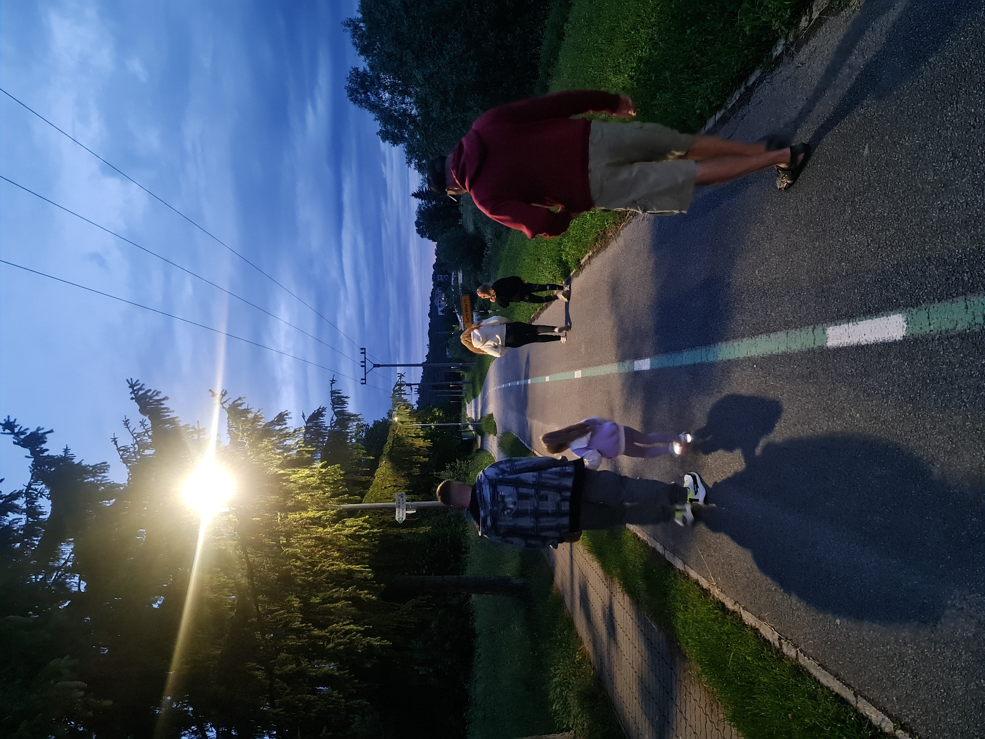 The image depicts a paved path, likely a bike path or walkway, in Prešov, Slovakia, during twilight. The path is marked with a green and white stripe. Four individuals are walking away from the camera on the path. On the left, a man wearing a patterned jacket and dark pants walks with a young girl in a purple hooded jacket and leggings. Further ahead on the path, two young women, one in a white hoodie and black pants, and the other in a black hoodie and leggings, walk together. To the right, a man in a maroon hoodie and khaki shorts walks ahead of the group.
The environment includes trees and vegetation lining the path. A street lamp on the left illuminates a section of the path and nearby trees. In the background, there are more trees, power lines supported by utility poles, and distant buildings. The sky is dark blue with clouds, indicating the transition from day to night. A road sign indicating "Euro Velo 11" is visible in the background, suggesting the path is part of a cycling route. The shadows of the people are cast long on the path, further confirming the low angle of the light.