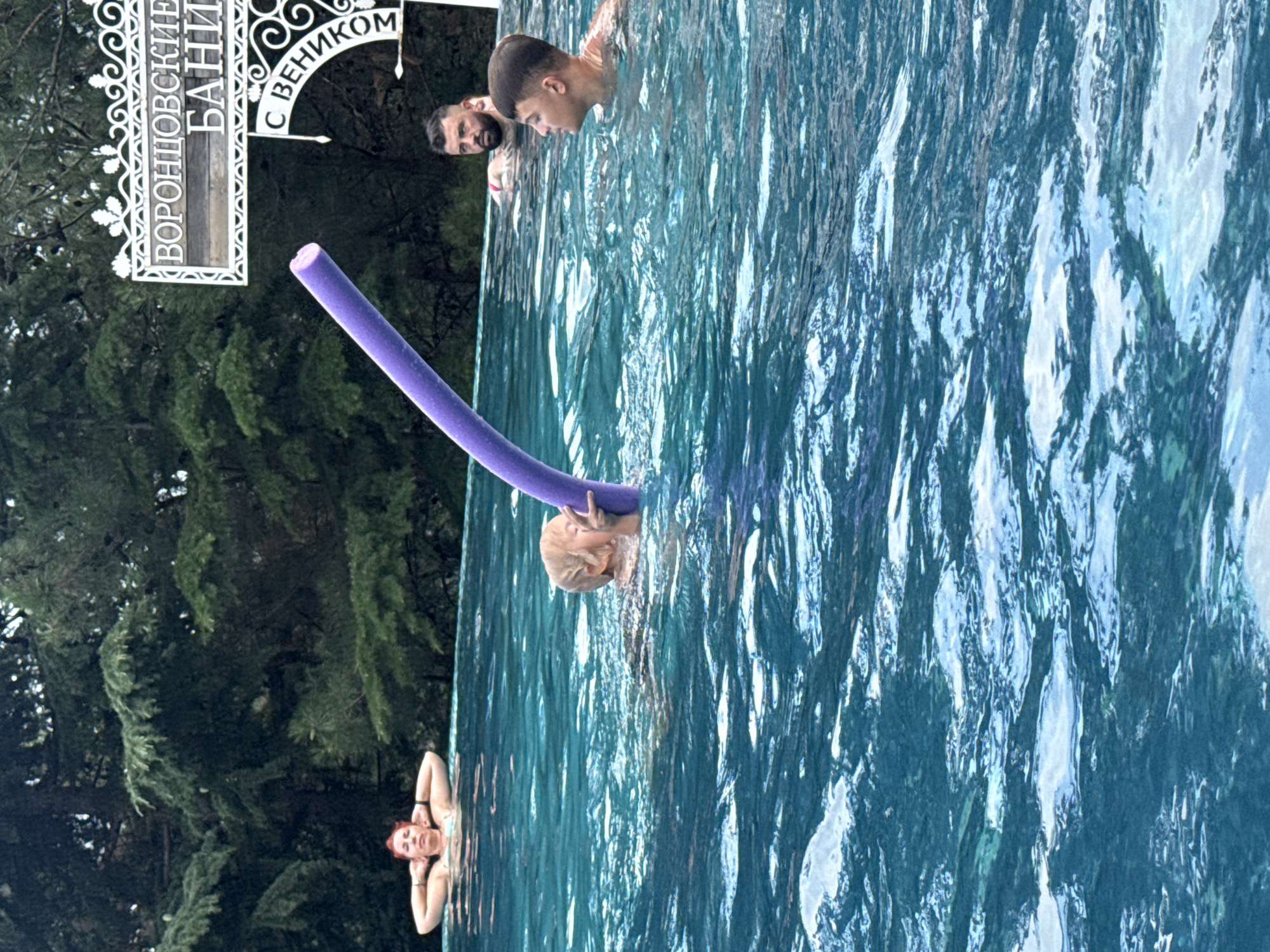 In the city of Yalta, Ukraine, four people are visible in an outdoor swimming pool. A woman with red hair is relaxing in the water near the edge of the pool, with her hands behind her head. In the middle of the pool, a child is holding a purple pool noodle and making a peace sign with their fingers. To the right of the child, two men are in the water. One man, with dark hair and a beard, is looking towards the left. The other man, who is younger and has short dark hair, is looking to the right. A sign with Cyrillic lettering is visible in the background, reading "ВОРОНЦОВСКИЕ БАНИ" and "С ВЕНИКОМ". Lush green trees surround the pool area.