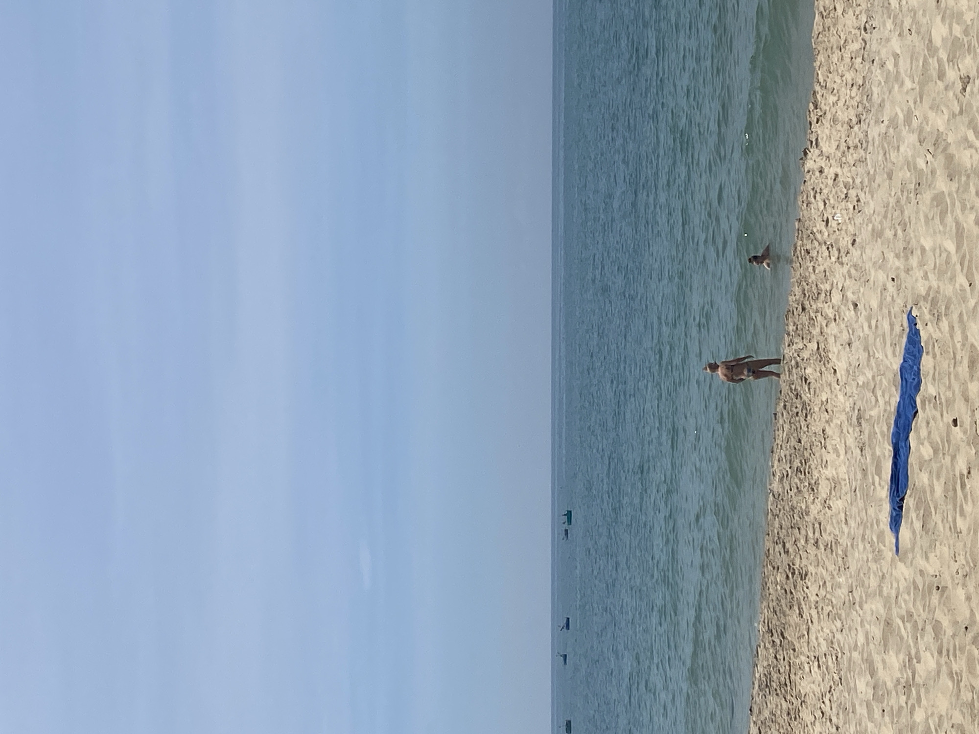 This is a photograph taken at the beach in Phu Thuy Ward, Vietnam. In the foreground, the sandy beach is visible with a blue towel laid out. In the middle ground, the ocean meets the shore, with gentle waves. Two individuals are in the water; one appears to be standing and facing away from the camera, while the other is partially submerged, with only their head visible. In the background, the calm sea stretches to the horizon, with several small boats visible in the distance under a hazy, pale blue sky. The overall impression is a serene beach scene on a clear day.
