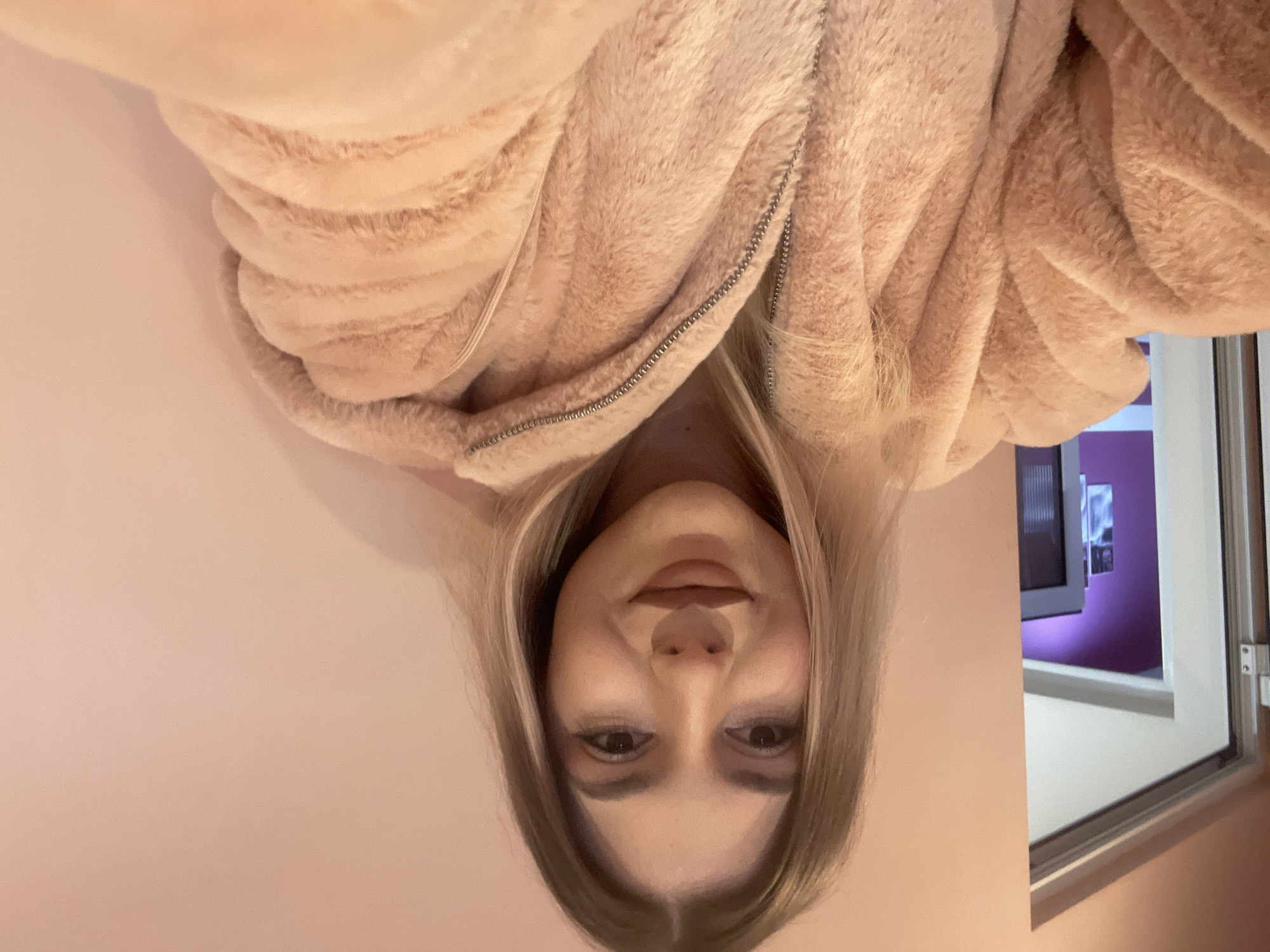 A woman is facing the camera in an upside-down orientation. She has fair skin, brown eyes, and light blonde hair that falls to her shoulders. She is wearing a soft, light pink fuzzy zip-up jacket. A window is visible in the background with white frames, revealing a purple-toned wall with hanging artwork. The background wall the woman is against is a light pink color. There is no information to identify the location in Burgas, Bulgaria, or any other location.