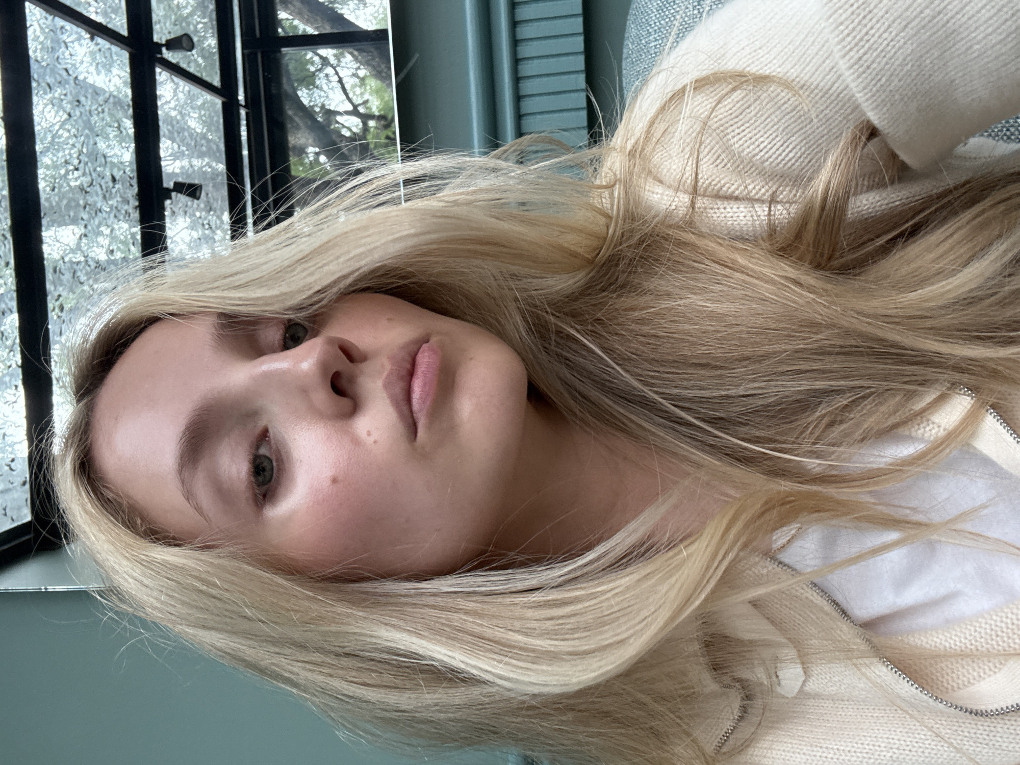 A young Caucasian woman with blonde hair and fair skin is captured in a close-up, front-facing portrait. She is wearing a cream-colored cardigan over a white top, both featuring textured knit patterns and a metallic zipper detail on the cardigan. The background suggests an indoor setting with a teal-colored wall to the left, featuring subtle horizontal paneling, and a glass ceiling with visible structural elements and tree branches through it in the upper right portion of the frame. The lighting appears to be natural, casting soft shadows and highlighting the woman's facial features, including her green eyes and light pink lips. A small mole is visible on her right cheek. The overall impression is one of casual intimacy and natural light. The presence of trees seen through the glass ceiling suggests a location with an outdoor view, possibly a restaurant, cafe, or a home with a conservatory-like feature. Given the context provided by the prompt, this scene is situated in Mexico City, Mexico.
