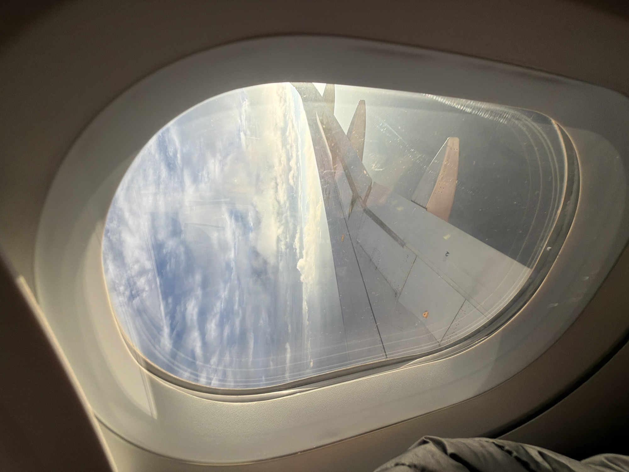 An aerial view from an airplane window shows a wing extending across the frame. The sky above is filled with clouds, some wispy and others denser, with sunlight breaking through. Below the clouds, the sea or land is visible as a dark expanse. The interior of the airplane cabin is visible around the window frame, with a hint of a passenger's clothing on the left side. There are no discernible landmarks or urban features that would indicate the city of Upshi, India, or any specific location.
