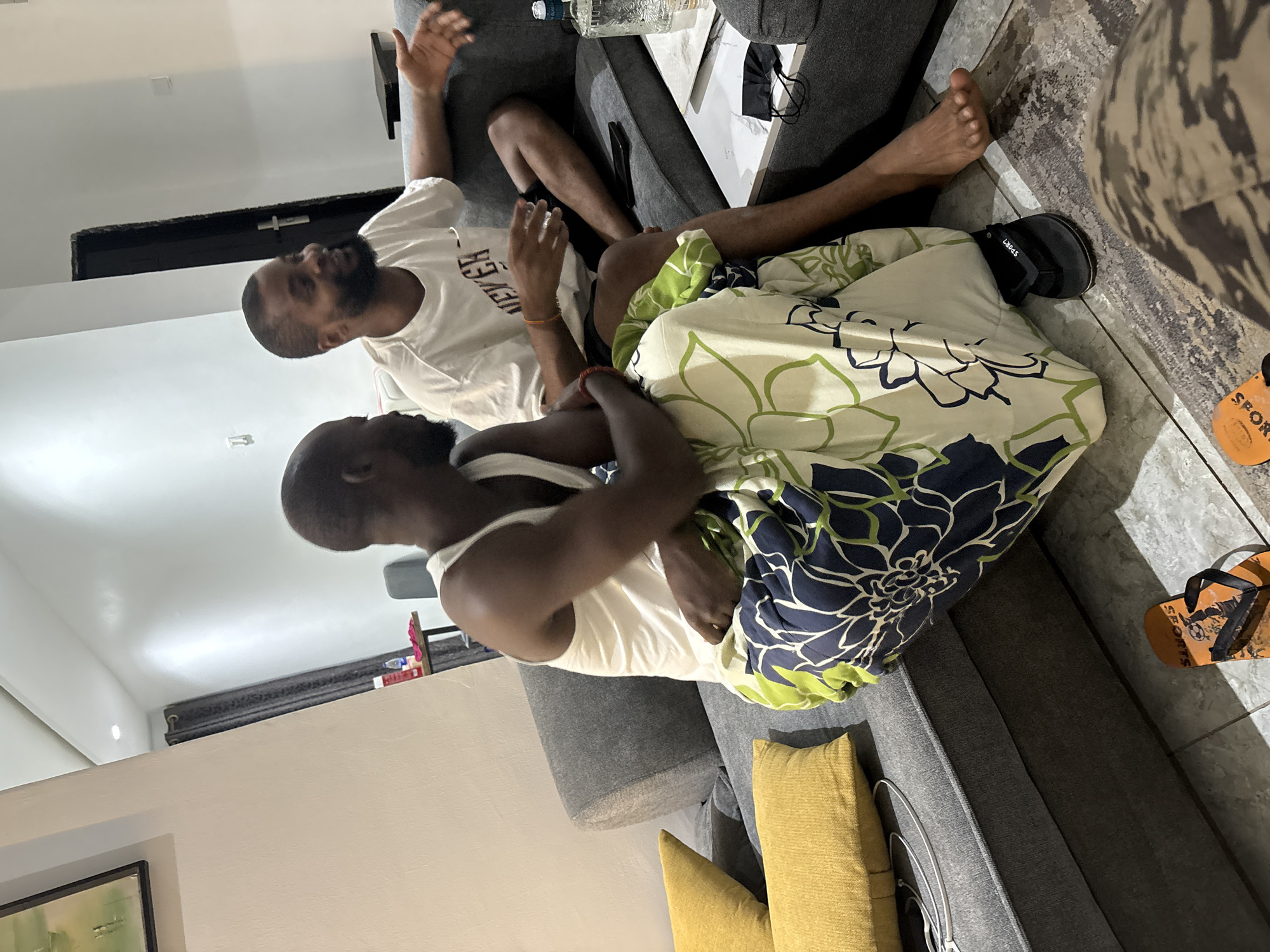Two men are casually seated on a grey sectional sofa in an indoor setting, likely a living room. The image is oriented vertically but depicts the scene horizontally.
The man on the left wears a white T-shirt, partially obscured, with the visible text "EVER" on it, and dark shorts. He leans back on the sofa, gesturing with his raised right arm as if speaking, and has a dark beard. The man on the right, also with a beard, wears a white tank top and is partially covered by a distinct floral-patterned blanket featuring green, white, and dark blue designs. He sits facing the first man, engaged in conversation.
Yellow throw pillows are on the sofa, and a clear glass bottle is visible on a white surface behind the sofa. The floor is covered with grey tiles. In the foreground, two flip-flops are visible on the tiled floor: one black with "SPORTS" written on the strap, and another orange one with "SPORT" visible. A dark door or cabinet is visible in the background against white walls. The indoor lighting suggests the scene takes place in the evening or at night. The atmosphere appears relaxed, and the men are engaged in a casual discussion. The attire and general setting are consistent with a warm climate like Iddo Tudunwada, Nigeria.