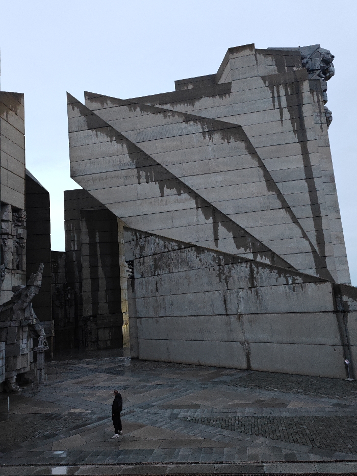 The image displays a forensic view of the Monument to 1300 Years of Bulgaria, located in Shumen, Bulgaria, under an overcast sky. The monumental structure is predominantly constructed from large, grey concrete blocks arranged in angular, Brutalist architectural forms. A prominent diagonal concrete wall with a series of stepped, pointed edges extends from the mid-left to the upper right of the frame, exhibiting numerous dark vertical water stains and streaks. To the left, a separate section of the monument incorporates large, blocky sculptural elements, including a partial view of a figure holding an elongated object, integrated into the concrete. A sculpted head, possibly zoomorphic, protrudes from the upper right section of the main diagonal wall. The ground in the foreground is paved with grey flagstones and cobblestones, appearing wet and reflective in areas, consistent with recent precipitation. One adult individual is present in the lower-mid foreground, standing centrally. This person is dressed in dark outerwear and light-colored shoes, facing towards the right side of the frame, observing the monument. No other individuals or dynamic actions are depicted.