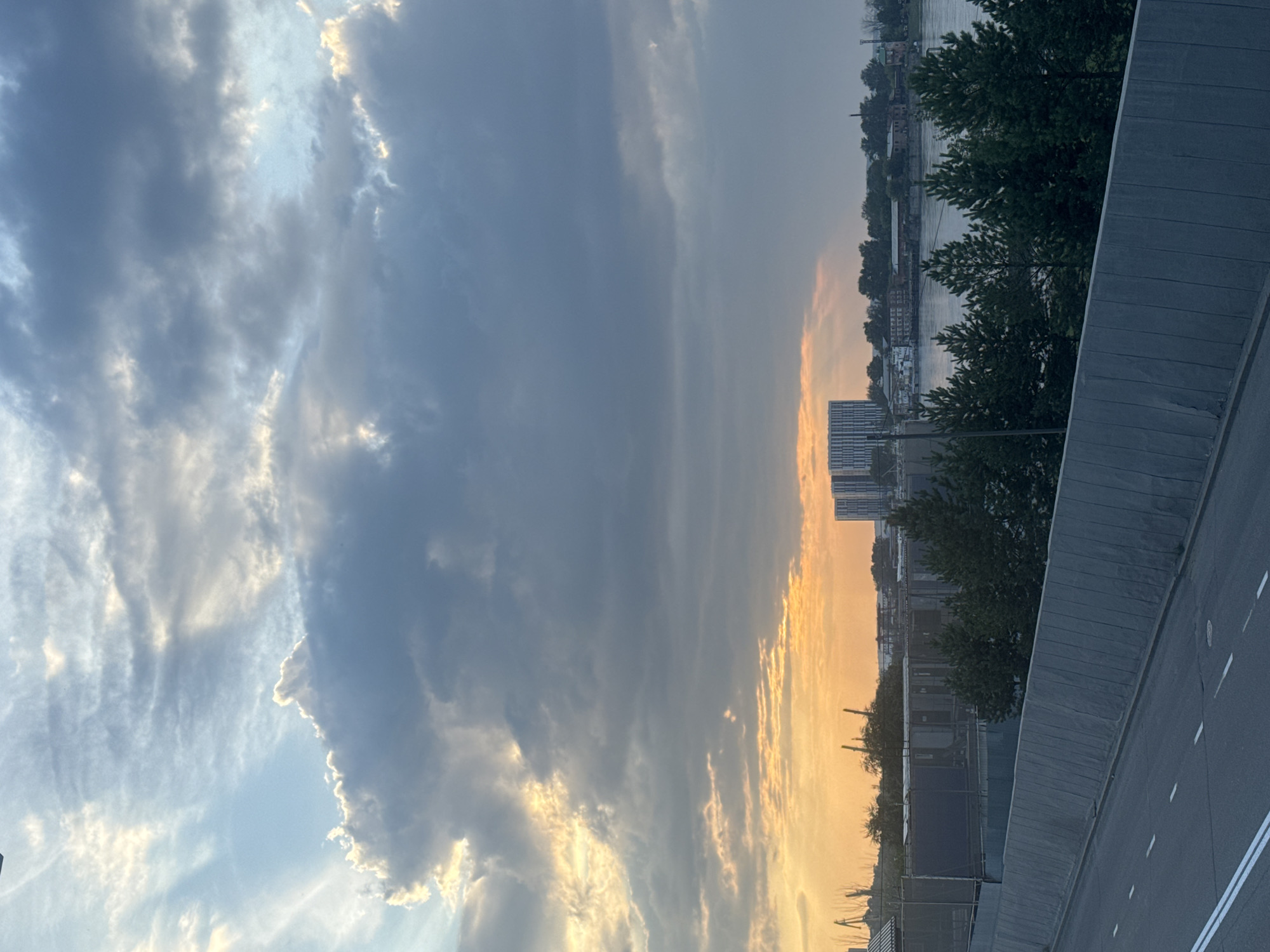 The image captures a dramatic sky with a mix of dark and light clouds, illuminated by streaks of sunlight. The setting appears to be during sunset or sunrise, given the warm orange glow at the horizon. In the middle ground, a wide river flows, bordered by trees and buildings on the far bank. Notably, a large, modern-looking building with a striped facade stands prominently on the left side of the far bank, while a cluster of older, industrial-style buildings with smokestacks are visible further to the right. A highway or road with white markings runs in the foreground, separated from the scene by a grey concrete barrier. The overall impression is one of a developing urban or industrial area situated near a body of water, with a striking natural sky. While the image does not explicitly identify the location, it resembles the industrial and riverside areas of Irkutsk, Russia, particularly areas along the Angara River where such architecture and infrastructure are common.