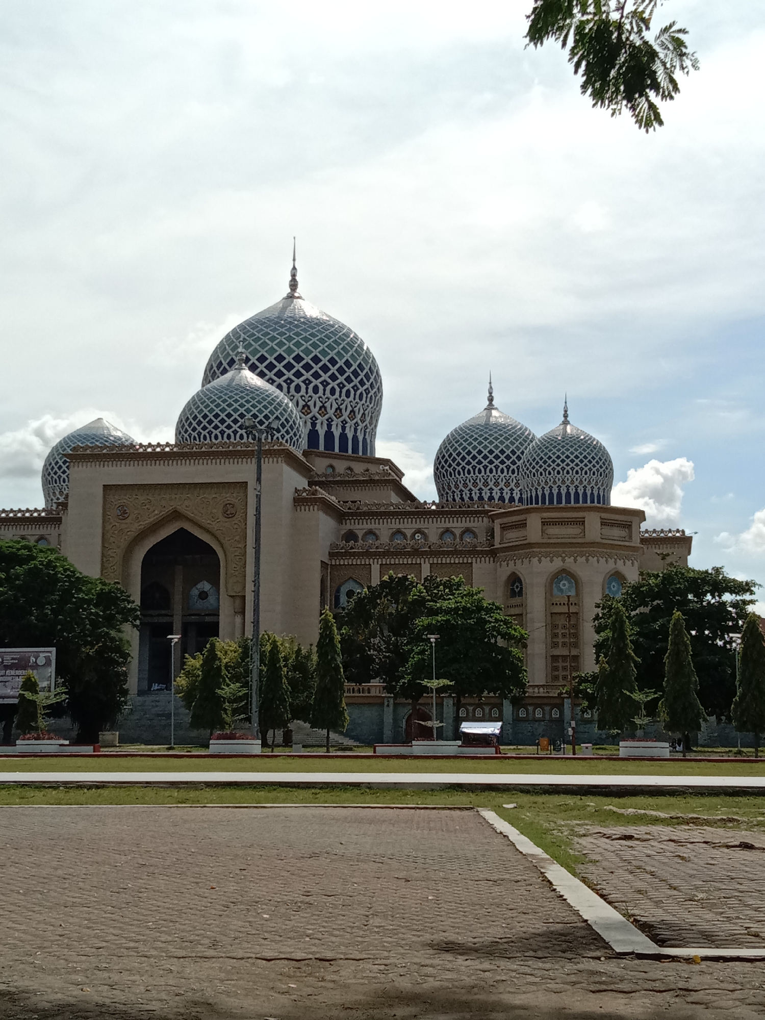 The image depicts the Islamic Center of Lhokseumawe, a religious and cultural complex located in the city of Lhokseumawe, Indonesia. The complex features a grand mosque with multiple domes covered in a lattice pattern in blue and green colors. The main building is made of light brown stone and decorated with intricate carvings. In the foreground is a paved courtyard area with a strip of green lawn bordering it. Several small, conical-shaped trees are planted in the median separating the mosque from the courtyard. A few benches can be seen in the vicinity of the trees. Some taller trees are also interspersed around the mosque grounds. The sky is partly cloudy with diffused sunlight. There are no visible people in the image. A corner of green foliage from a tree is visible in the upper right corner.