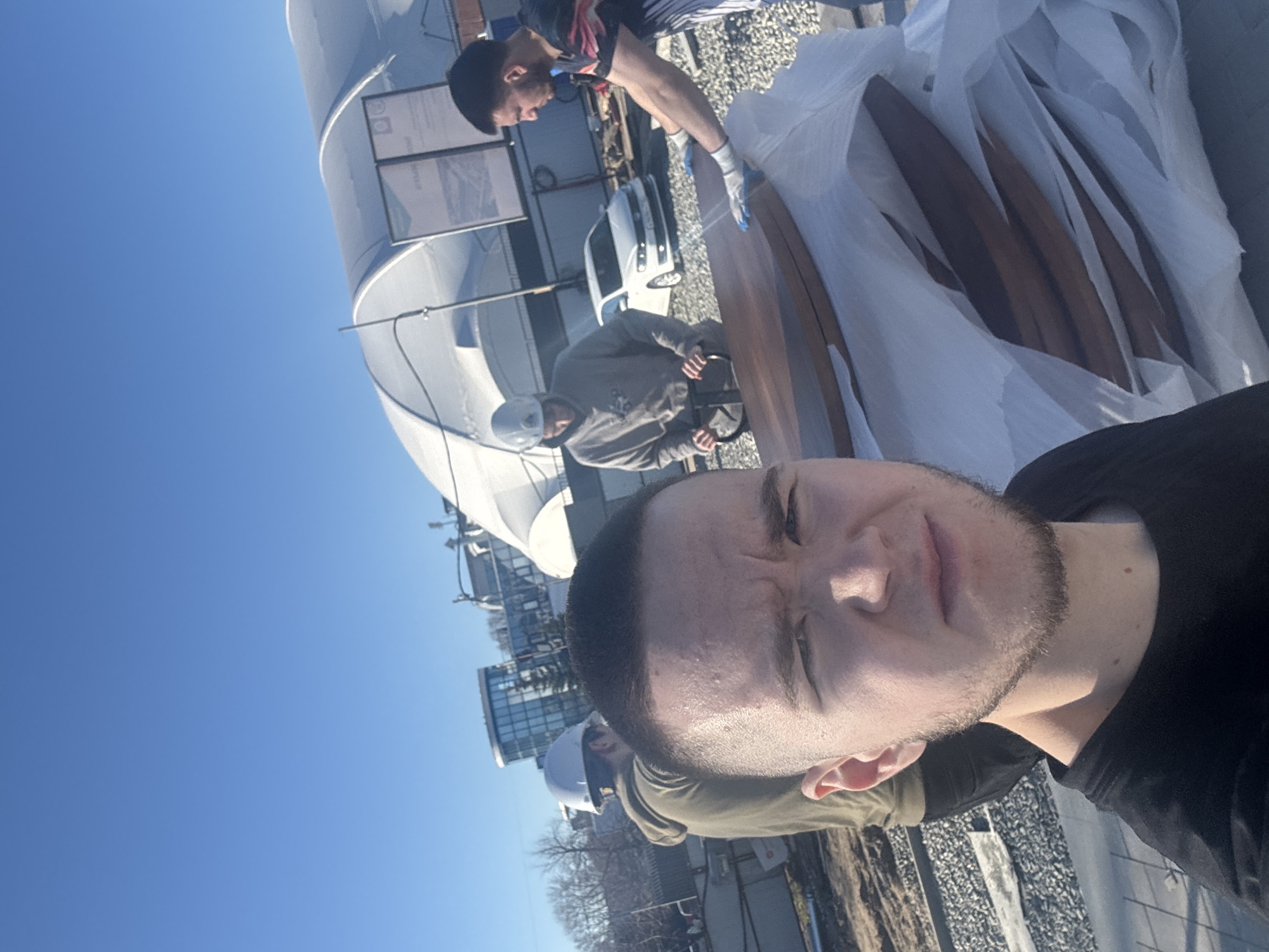 The image depicts a selfie taken on a rooftop in Kazan, Russia. In the foreground is a fair-skinned man with dark hair and a beard, wearing a black shirt. Behind him, two men are working with wooden boards, partially covered in white plastic. One man is wearing a helmet and a gray shirt, and the other is wearing a patterned shirt and blue gloves. A white van is parked nearby. In the background, a large white cylindrical structure is visible, along with buildings and a clear blue sky. The image is slightly rotated, creating a tilted perspective.