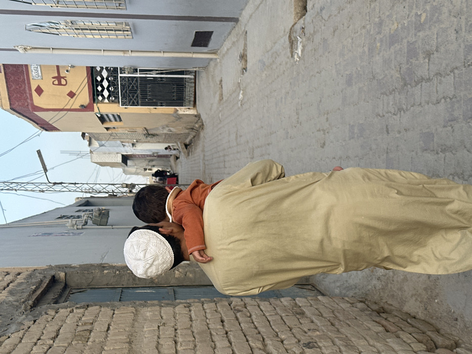 An adult male, wearing a light tan long-sleeved tunic and a white crocheted skullcap, is observed from behind, walking away from the camera down a paved residential alleyway in Quetta City Tehsil, Pakistan. He is carrying a young child, dressed in an orange long-sleeved top, on his left hip/shoulder. The child's head is turned away from the viewer. The alley floor is constructed of grey interlocking concrete pavers, showing signs of wear and minor surface irregularities.
To the immediate left, a textured, unpainted brick wall is visible, with a light blue door set within it further down. To the immediate right, a light grey building with two vertically oriented arched windows featuring metal grilles is present. Beyond this, a light yellow building with a maroon roofline trim and a black metal entry gate is visible. Decorative patterns, including a stylized crescent or 'U' shape and Arabic script, are painted on the yellow building's facade.
In the middle distance, centrally aligned with the alley, a tall metal lattice utility structure (possibly an electricity or communication pylon) supports numerous black overhead wires that crisscross the street. A single, simple streetlight fixture is attached to this structure. Further down the alley, faint figures are discernible, including what appears to be an adult figure near a red object (possibly a handcart) and a smaller figure seated or standing near a building. The sky is a hazy, light blue, indicating daylight conditions.