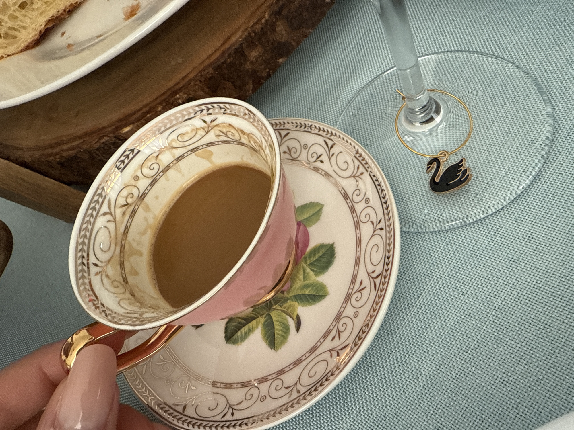 The image shows a tea cup, partially filled with a light-colored liquid, resting on a matching saucer. The cup is predominantly pink with gold trim, while the saucer features a floral motif and gold trim. A wine glass with a gold ring and a black swan charm is positioned next to the saucer. A person's hand, with long, light-colored nails, is partially visible holding the teacup. The cup and saucer are placed on a light blue, textured surface, likely a tablecloth. A wooden tray with a white plate and some food is located in the upper left corner of the image. The location is Davie, United States of America.